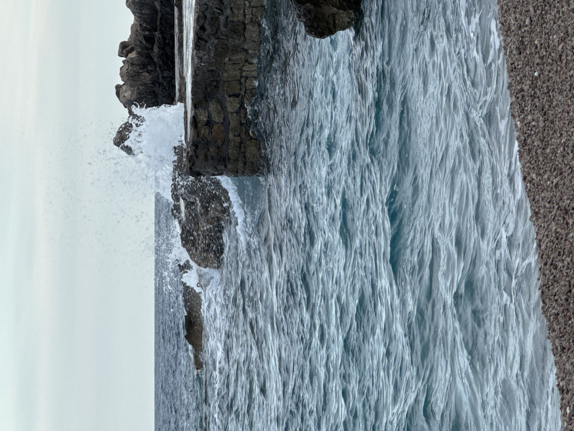 An action shot captures a wave crashing against a rocky shore and a stone seawall in Prijevor, Montenegro. The rough sea is a deep blue with white caps, and the wave is frozen in mid-air as it explodes against the dark rocks and the weathered stone structure. The sky above is a pale, overcast gray. The foreground is a gravelly beach. The image focuses on the raw power of the sea against the man-made and natural elements of the coastline.