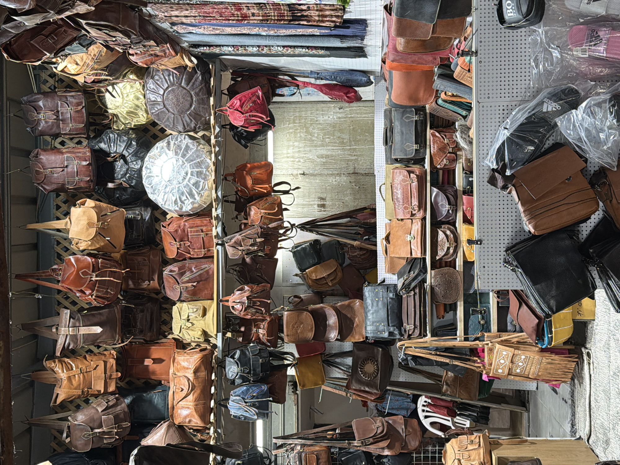 This media file captures a densely packed market stall or shop, showcasing a vast array of leather goods, primarily bags. The scene is filled with numerous leather bags of various sizes, styles, and shades, ranging from light tan to dark brown and black, along with some red/maroon items. Many bags feature prominent buckles, straps, and flap closures, suggesting messenger bags, satchels, and backpacks.
Interspersed among the bags are large, round leather poufs or ottomans, including one dark brown with an intricate embossed design, a shiny gold one, and a glossy silver one. Above and behind the leather goods, folded textiles such as scarves or blankets are stacked on shelves. On the right side, an assortment of black footwear, possibly sandals or slippers, are visible in clear plastic packaging. Several folded umbrellas or parasols are also displayed.
The goods are displayed in a rustic manner, hanging from hooks, arranged on shelves, and stacked, giving the impression of an authentic marketplace. The overall lighting suggests an indoor or covered outdoor setting, possibly during the daytime. While no people are visible, the dense display indicates a retail environment.
Visible text includes "BOSS" on a black cap or bag towards the top right of the original image, and "adidas" on the packaging of the black slippers/sandals. The location context provided is Tel Aviv, Israel, which aligns with the market aesthetic.