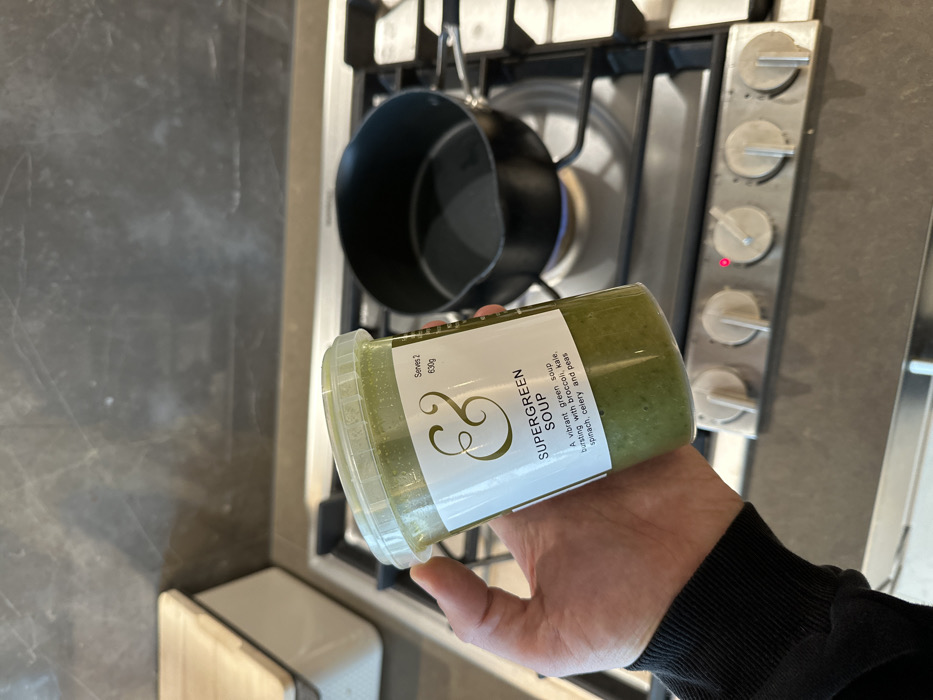 A human left hand, wearing a dark long-sleeved garment, holds a clear plastic container of "Supergreen Soup" in a kitchen. The container's label displays a golden ampersand logo, the product name "SUPERGREEN SOUP," ingredients listed as "broccoli, kale, spinach, celery and peas," "Serves 2," and "630g." The soup inside is a vibrant green. Directly beneath the container, a small, dark saucepan rests on the front-left burner of a stainless steel gas hob. This burner is actively lit, emitting a blue flame. The corresponding control knob on the hob's front panel displays a small illuminated red indicator light. The background features a dark grey mottled backsplash. To the far left, a light-colored wooden item and a white object are partially visible on a countertop. The scene is located in a residential kitchen in London, United Kingdom.