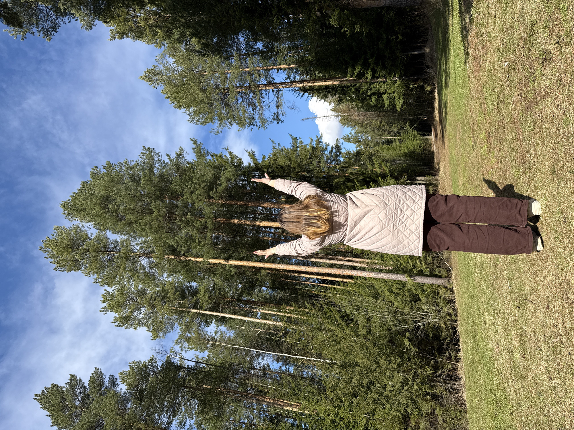 A woman stands with outstretched arms in a forest or park. The location is likely in or near the city of Адам, Russia. She wears a beige quilted jacket and brown pants. Tall trees rise towards a blue sky with scattered clouds. The ground is covered with grass and patches of dry vegetation.