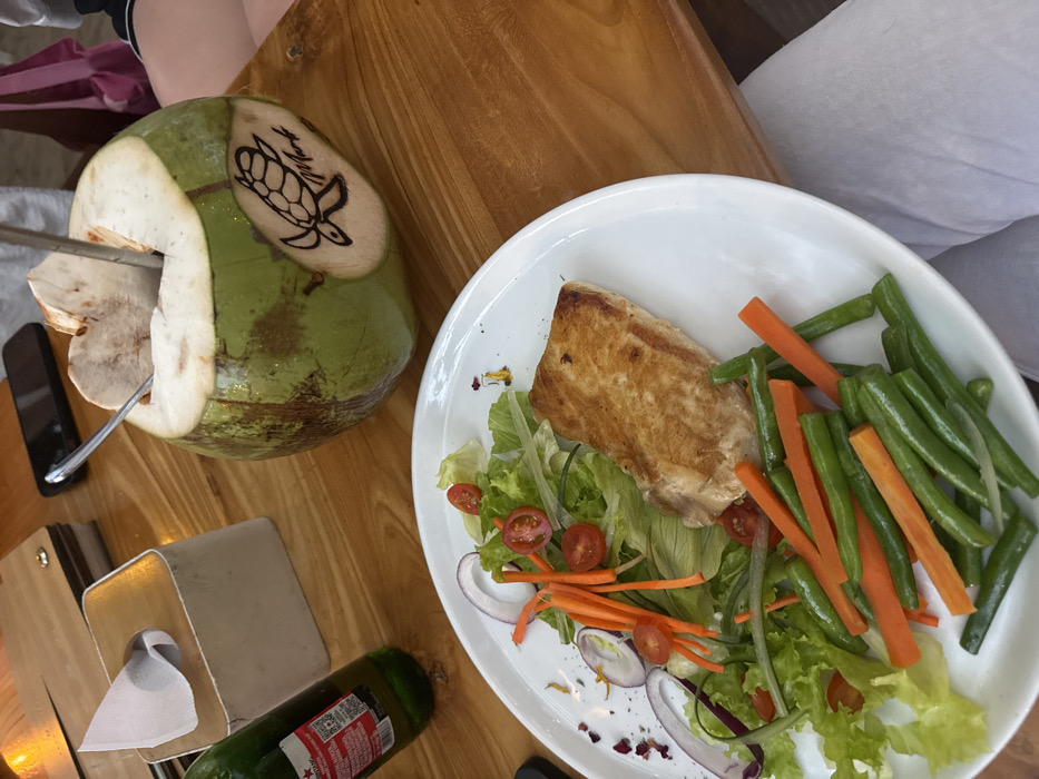 The scene is located in a dining establishment in Ped, Indonesia. The image depicts a meal set on a light-colored wooden table with visible grain. In the immediate foreground, a white oval plate holds a cooked rectangular cut of protein, accompanied by a green salad consisting of lettuce, sliced red onions, cherry tomatoes, and shredded carrots. A separate portion of green beans and julienned carrots is arranged on the same plate. To the right of the plate, a green whole coconut, opened at the top with a metal straw inserted, rests on the table. The coconut displays a carved or burned depiction of a sea turtle and the textual inscription "Murk" on its side. To the left, a green glass bottle with a partially visible red and white label stands next to a light-colored, rectangular tissue box. A dark-colored smartphone lies flat on the table behind these items. The lower right corner reveals the upper legs of an individual wearing white trousers. A segment of a pink bag or fabric is visible in the upper right background.