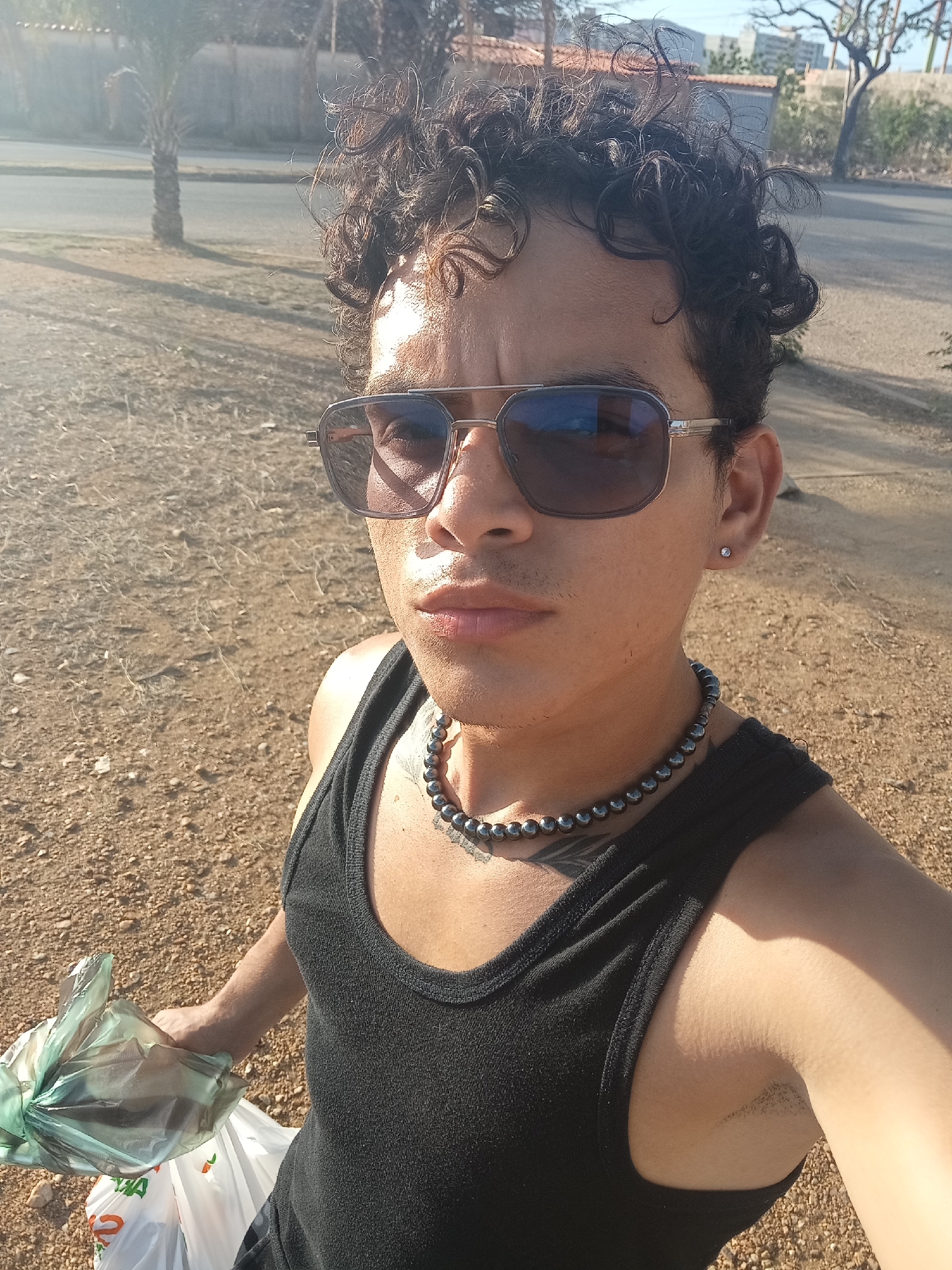 A young adult with curly dark hair and sunglasses is taking a selfie outdoors. They are wearing a black tank top and a beaded necklace. They are holding two plastic bags, one clear green and the other white with orange writing. The background shows a dirt path, a road with a palm tree and some buildings in the distance. The lighting suggests it is daytime. This location is identified as Juan Griego, Venezuela.