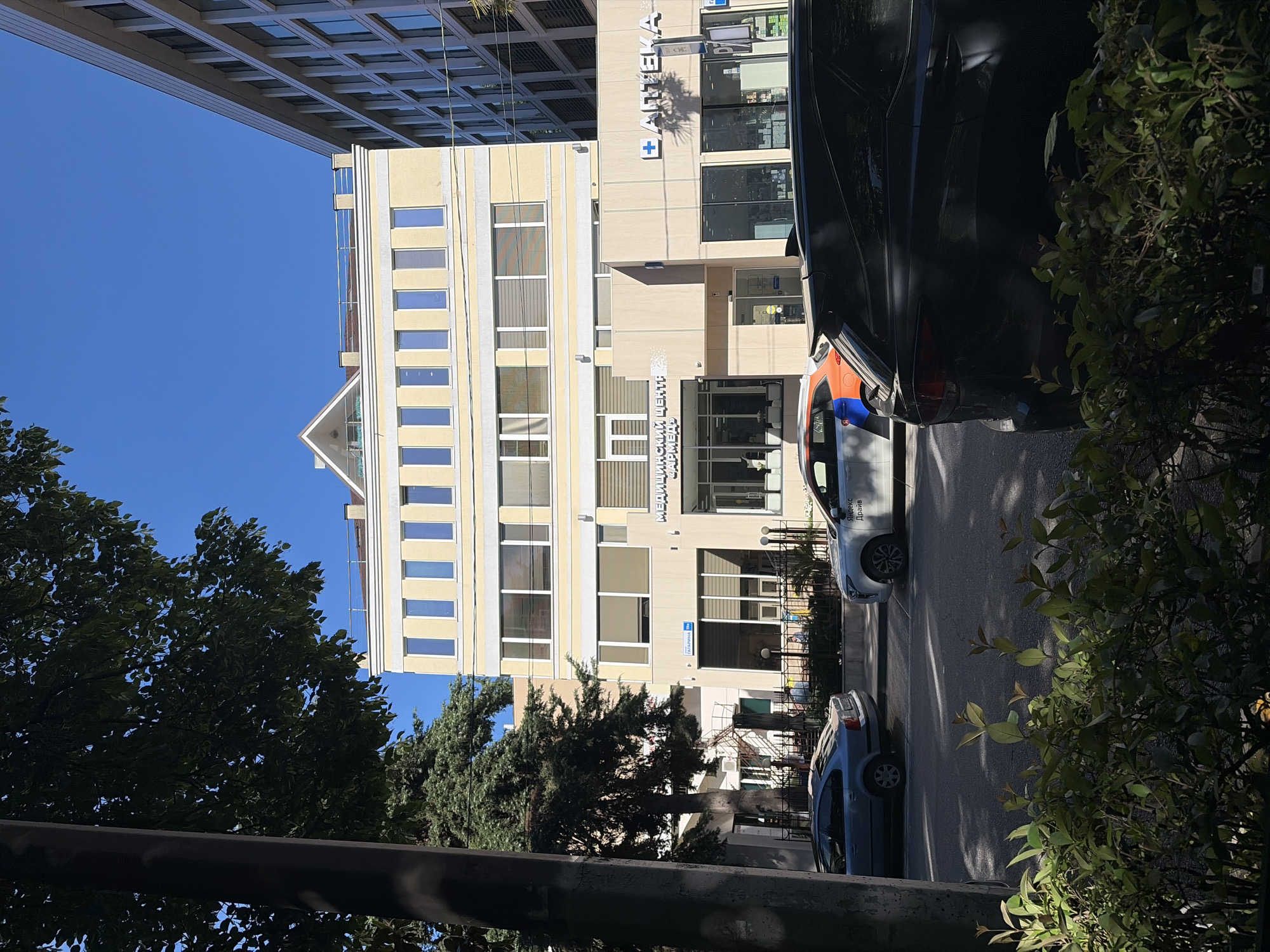 The image captures an urban street scene in Sochi, Russia, on a bright, clear day, likely during morning or afternoon due to the strong sunlight and defined shadows. The primary subject is a multi-story, light-colored (cream/yellow) commercial building with numerous windows.
On the facade of this building, prominently displayed, is the word "АПТЕКА" (APTEKA), meaning "Pharmacy," accompanied by a blue cross logo. Below this, further text reads "МЕДИЦИНСКИЙ ЦЕНТР ЗАРИМЕД" (MEDITSINSKIY TSENTR ZARIMED), indicating a "Medical Center Zarimed." A street sign with the name "ГЛАДИНА" (GLADINA) is also visible near the building's entrance.
To the left of this main building, a taller, darker, modern building with a distinct grid-patterned facade can be seen, partially obscured by the clear blue sky.
The street itself runs in the foreground, with several vehicles parked alongside. A white car with "Яндекс Драйв" (Yandex Drive) branding is clearly visible, suggesting a car-sharing service. Another car, partially visible, has distinctive orange and blue markings.
The scene is framed by lush green trees and bushes on both the left and right sides, adding natural elements to the urban setting. No specific activity or people are discernible, presenting a static snapshot of a city street. The weather is sunny with a clear blue sky.