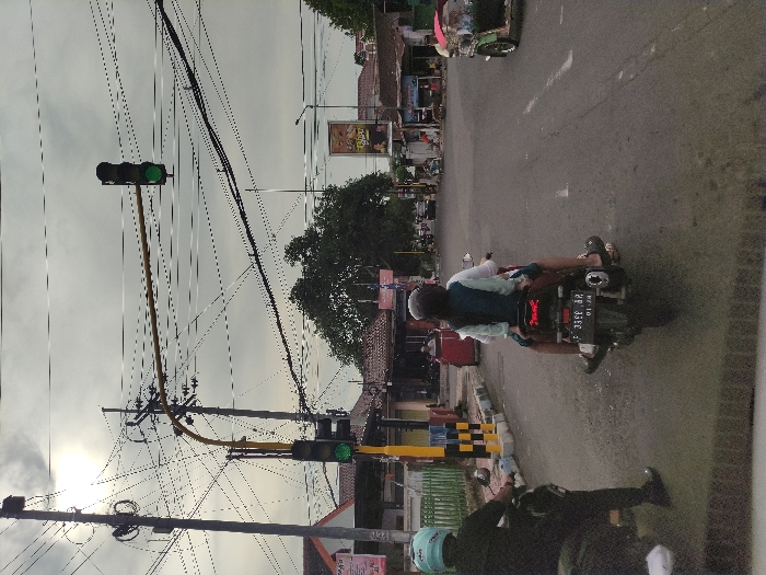 An outdoor street scene in Bondowoso, Indonesia, depicts traffic moving under an overcast sky. The image focuses on a road lined with buildings and utility poles, densely crisscrossed by overhead wires.
In the mid-ground, a person wearing a light-colored shirt or jacket and a helmet rides a dark motorbike away from the viewer. A blue backpack or vest is visible on their back. The motorbike has a license plate displaying "P 3356 JP" with "01 28" below it. The rider's feet, wearing sandals, are visible on the bike's footrests. Further down the road, a green traffic light hangs above the street, indicating that traffic in this direction is permitted to proceed. Another traffic light, also green, is partially visible in the background.
In the foreground, another person on a darker motorbike, wearing a light blue helmet and dark clothing, is partially visible on the left side of the frame. To the right of this motorbike, a yellow and black striped barrier or box is positioned by the roadside.
Low-rise buildings with shopfronts line both sides of the street in the background, with a billboard or sign visible above them on the right. A small tree stands in the mid-ground. The road itself is asphalt, with some faded markings. A tricycle or auto-rickshaw is partially visible in the far background, moving away from the camera. The sky is uniformly cloudy, suggesting either an early morning, late afternoon, or generally overcast day.