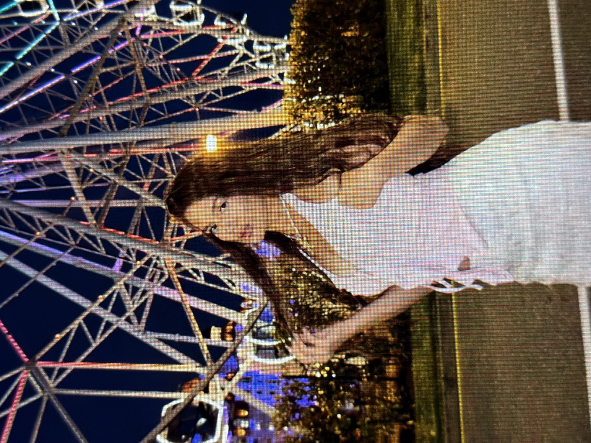 A young woman with long, brown hair is posed in front of a brightly lit Ferris wheel at night in the city of Sirius, Russia. She is wearing a light pink, halter-neck top and a white, sparkly skirt. Her left arm is raised, and her hand is playing with her hair, which cascades over her shoulder. The Ferris wheel, with its geometric structure and colorful lights, dominates the background. In the mid-ground, there are illuminated trees and bushes, and a yellow line runs horizontally across the image, suggesting a road or pathway. The overall atmosphere is festive and bright, illuminated by the artificial lights of the amusement park and the city.
