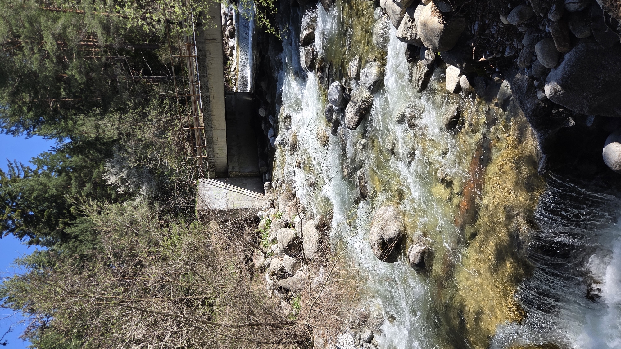 The image shows a flowing river with a concrete structure crossing over it. Large rocks line the riverbed and banks, with water cascading over them. Trees and other vegetation are visible along the river's edge, with a clear blue sky above the treetops. The location is Unknown, Unknown.