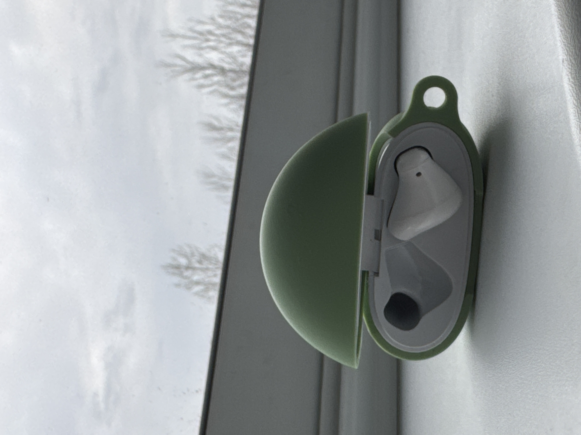 An eye-level shot captures a pair of white wireless earbuds nestled inside a light green, open protective case. The case, appearing to be made of silicone, features a circular loop at its top. One earbud is clearly visible within its compartment, while the second is partially obscured. The open case is positioned on a white or light-colored surface, likely a window sill or ledge, adjacent to a window.
Through the window, a blurred outdoor scene is visible. The sky is overcast and bright, suggesting a cloudy daytime. Bare tree branches, characteristic of a cold season like winter or early spring, are discernible against the soft grey backdrop of the sky. The lighting in the image is diffused and even. The composition is rotated 90 degrees counter-clockwise, causing the window and horizon to appear vertical. No visible text is present in the image. The setting depicts an indoor space looking out onto the environment of Ново-Никольское, Russia.