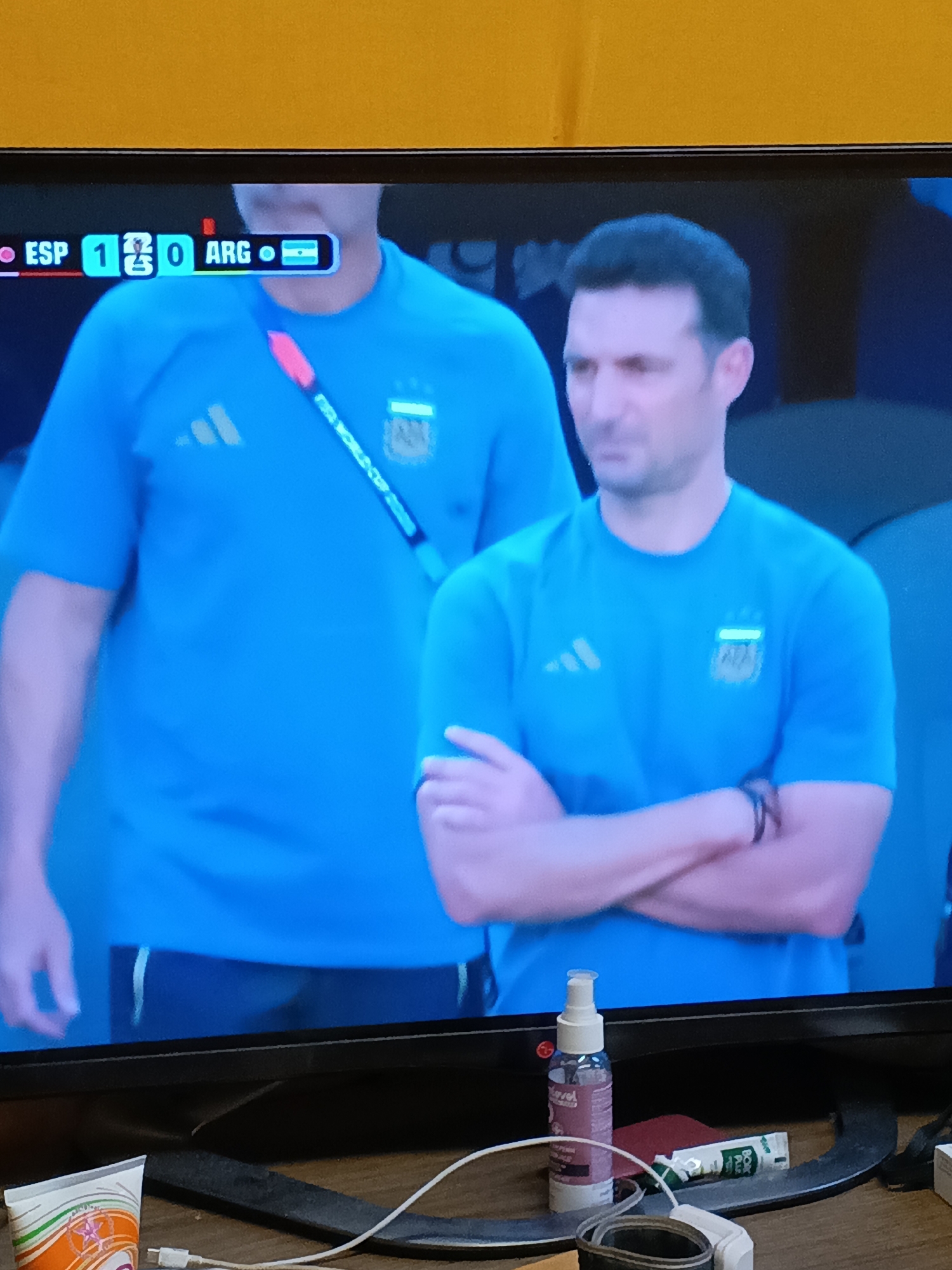 The image shows a television screen displaying a soccer match score with "ESP 1 - 0 ARG". Two men in blue soccer jerseys are visible on screen, with the man on the right in focus. He has dark hair and is looking to the left with his arms crossed. The background on the screen is a blurred soccer stadium. Below the screen, on a wooden surface, there are various objects including a bottle of hand sanitizer, a tube of balm, and a white cable. The top of the image has an orange background, possibly indicating the wall or ceiling above the television. The scene is likely indoors, as suggested by the lighting and the presence of the wooden surface and objects at the bottom. The location of Tatysh, Russia is not depicted or implied by any visible elements in the image.