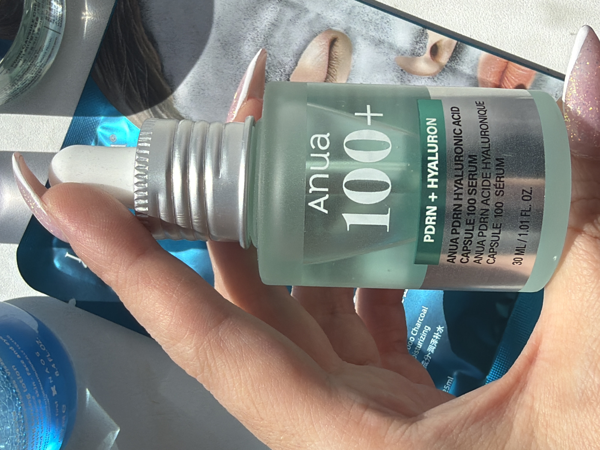 The image shows a hand holding a bottle of Anua 100+ PDRN + Hyaluron serum. The bottle is light green with white lettering. The product claims to contain Anua PDRN Hyaluronic Acid and is labeled as a Capsule 100 Serum. The bottle contains 30 ml / 1.01 fl oz of the product. The hand has long, manicured nails with glitter accents. The background is blurry but appears to contain other skincare products and a magazine or leaflet with an image of feet on it. The location is not identifiable as Saint Petersburg, Russia, as there are no clear indicators within the image.