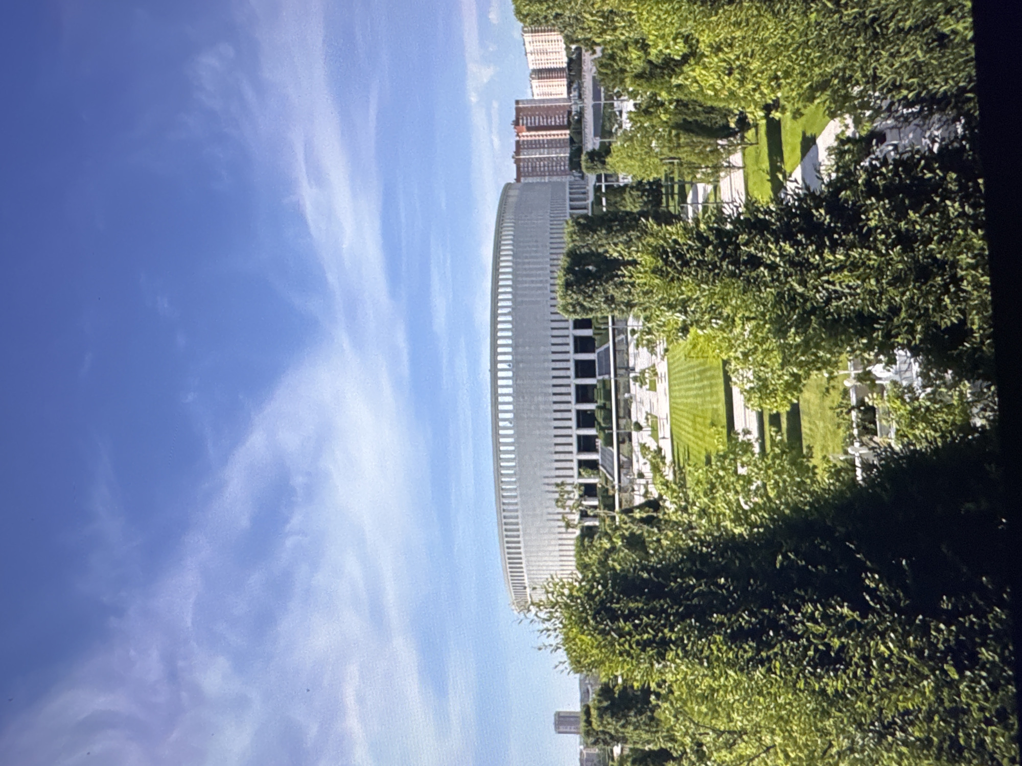 The image displays a modern, circular building with a white facade and numerous vertical slats, suggesting a sleek, contemporary architectural design. The building is surrounded by lush green trees and manicured lawns, indicating an urban park or a landscaped area around a corporate or institutional facility. In the background, several tall brick buildings rise against a clear blue sky with wispy white clouds, hinting at a cityscape. The lighting suggests it is daytime, possibly late morning or early afternoon, given the bright sunlight and clear visibility. The overall scene evokes a sense of calm and order, with a blend of natural elements and man-made structures. No people or specific activities are visible, focusing instead on the architecture and landscape. The image appears to be taken from an elevated perspective, possibly from another building or a slightly elevated vantage point. There is no visible text in the image.