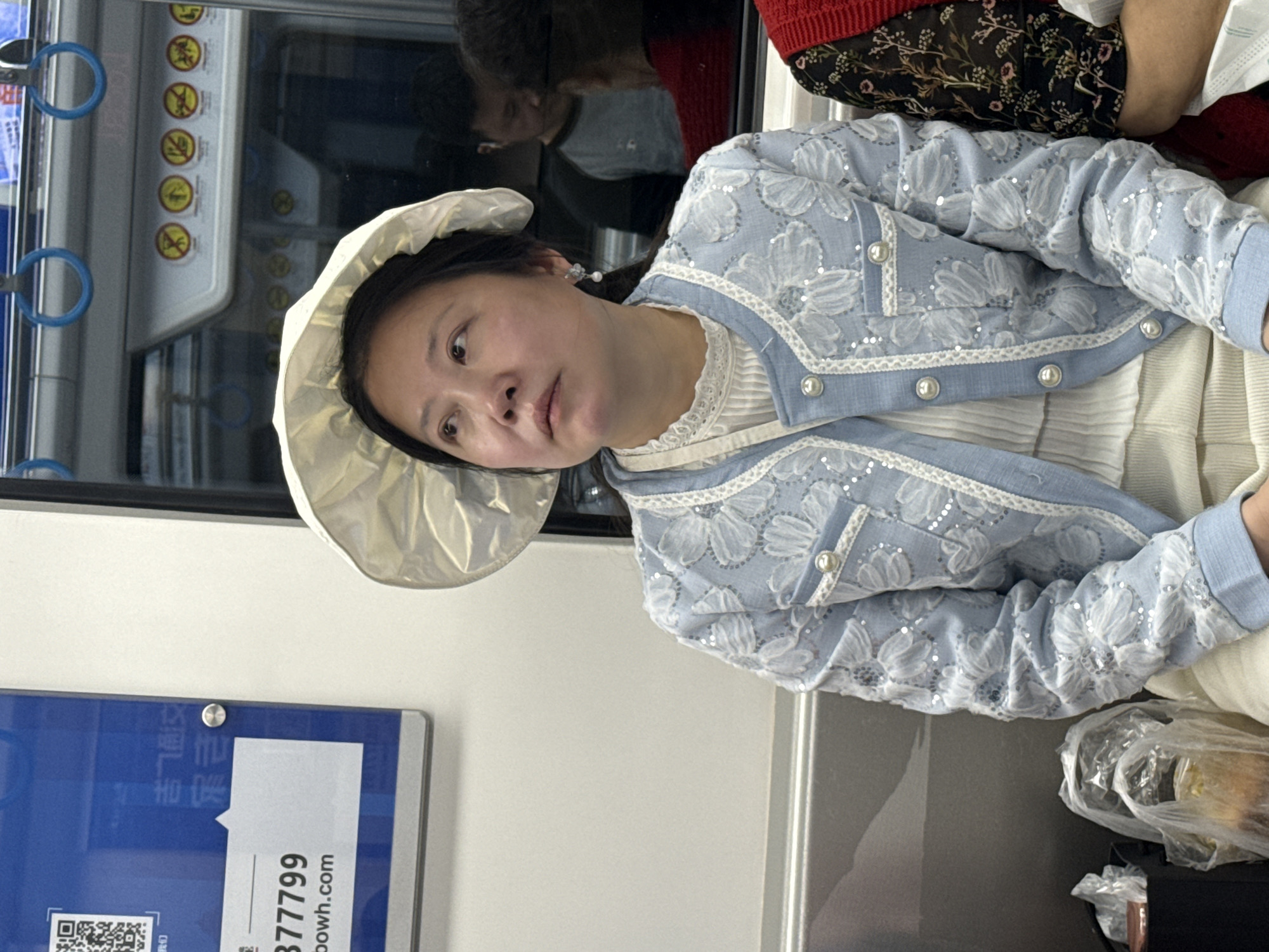 A woman wearing a wide-brimmed hat and a light blue jacket with white floral embroidery is seated on a subway train in Jiulongpo District, China. Another passenger can be seen behind her, reflected in the train window. A blue and white informational panel is attached to the train wall to the left of the woman. A plastic bag is partially visible in the lower right corner of the image.