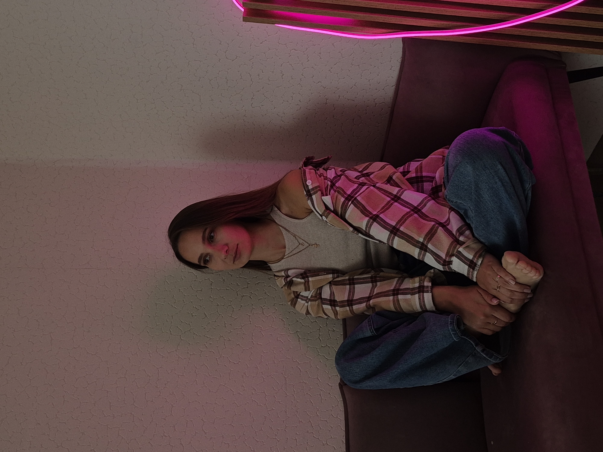 A young adult female is seated cross-legged on a dark maroon or plum-colored sofa in an indoor setting. She has long, straight brown hair and is looking towards the left side of the frame with a neutral, contemplative expression. She wears a light-colored ribbed tank top, an open plaid button-up shirt in shades of tan, brown, and pink, and loose-fitting blue jeans. Her bare feet are visible, with her right hand resting gently on her left foot. She has small rings on her fingers and toes, and two delicate necklaces, one featuring a small cross pendant. The background features a textured, light-colored wall with an uneven surface, illuminated by a strong pink/magenta glow emanating from an artificial light source on the right. This pink light casts distinct hues onto the wall, the woman's clothing, and the sofa. A portion of a vertical wooden slatted structure, integrated with a bright pink neon or LED light strip, is visible on the far right. The overall lighting is dim, emphasizing the pink illumination.
