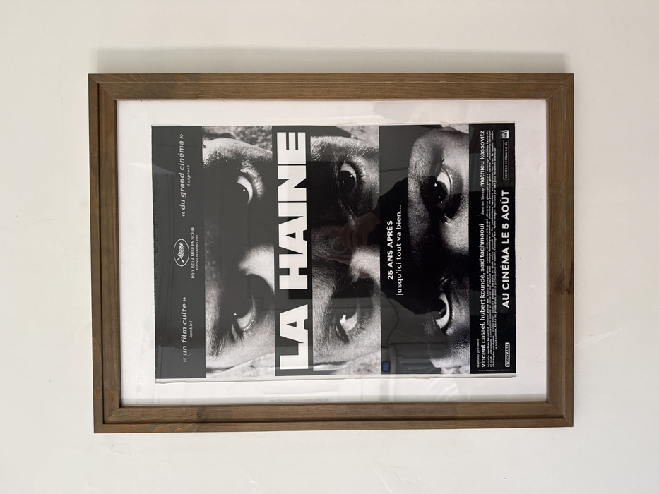 A framed movie poster for "LA HAINE" is displayed vertically on a plain white wall in an indoor setting within the city of New York, United States of America. The poster is encased in a brown wooden frame with a white mat board. The artwork is black and white, featuring three stacked horizontal segments, each depicting the eyes and forehead of a different man. The title "LA HAINE" is prominently centered in large white capital letters on a black band between the top two eye segments. Below the middle set of eyes, white text reads "25 ANS APRÈS jusqu'ici tout va bien...". Additional white text at the top references "un film culte", "PRIX DE LA MISE EN SCÈNE FESTIVAL DE CANNES 1995", and "du grand cinéma". The bottom section lists actors Vincent Cassel, Hubert Koundé, Saïd Taghmaoui, and director Mathieu Kassovitz, concluding with "AU CINÉMA LE 5 AOÛT". A faint reflection of a person holding a camera or phone is visible across the poster's protective glass surface.