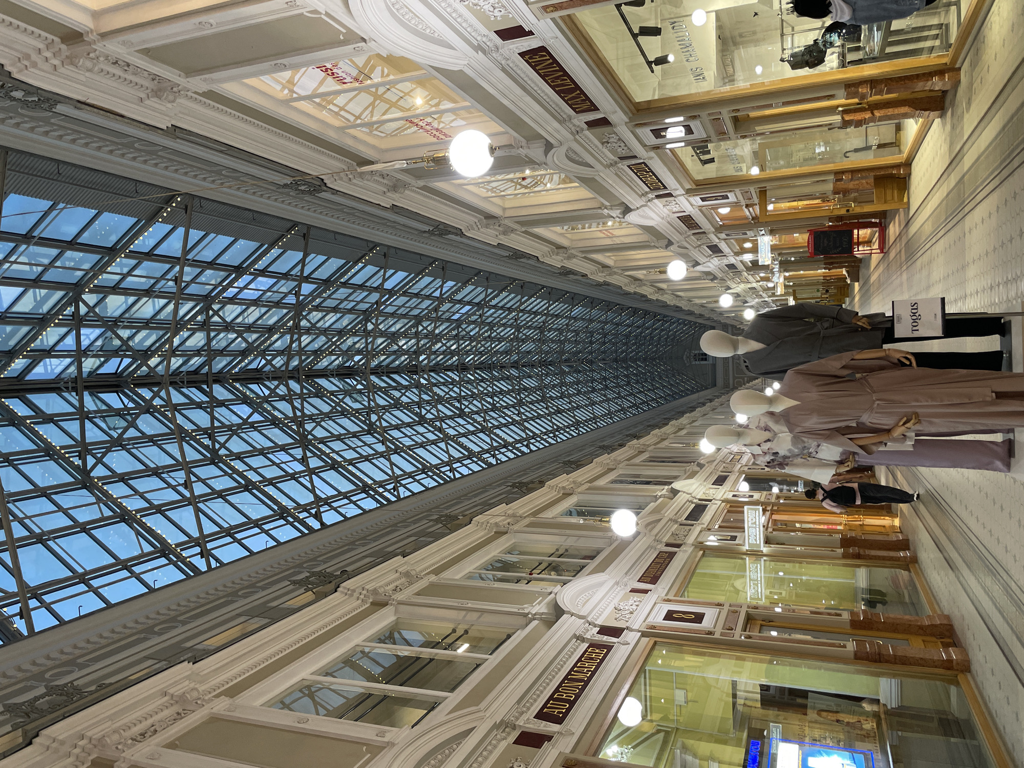 The image shows the interior of a shopping arcade in Saint Petersburg, Russia. The arcade features a large glass ceiling supported by a metal grid structure. Below the ceiling are decorative architectural details and signage with text, including "AU BON MARCHE". Mannequins wearing clothing are displayed in front of store windows. Lighting fixtures are visible, hanging from the ceiling. The arcade appears long and narrow, with the perspective drawing the eye towards the far end. A person is visible looking into the windows of a store.