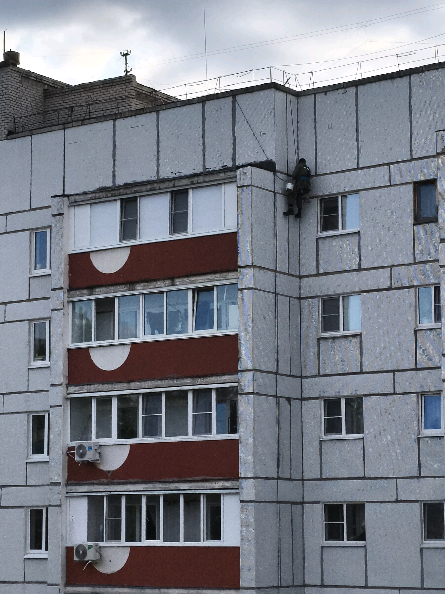 A single individual is performing maintenance work on the exterior of a multi-story apartment building. The person is suspended by ropes and appears to be working on the upper facade of the building, near the roofline. The building itself is constructed with grey concrete panels and features multiple rows of windows and balconies. Red decorative panels are visible on some of the balconies. The sky is overcast, suggesting a cloudy day. The scene is located in the city of Chaikovsky, Russia.