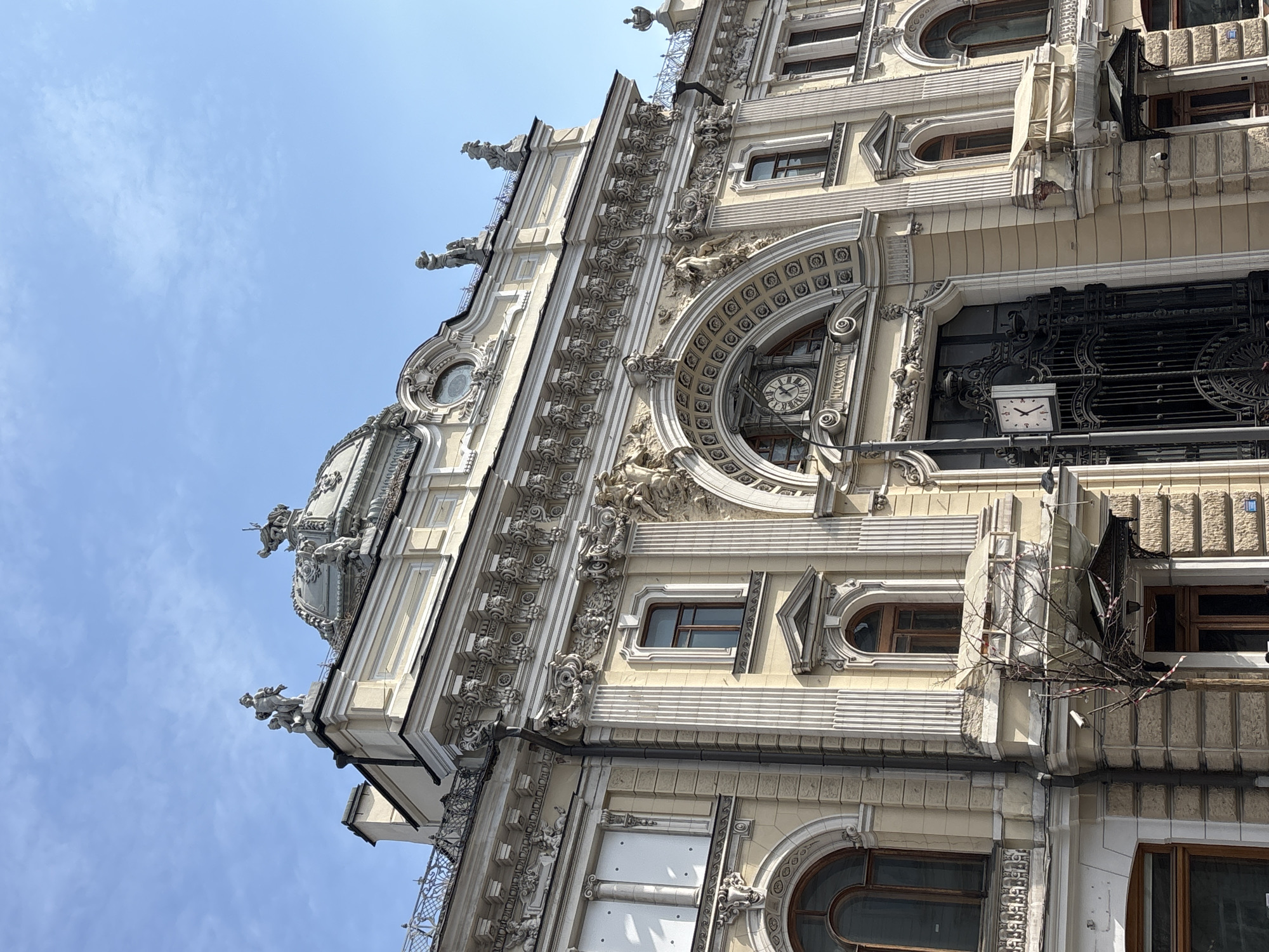 The image shows the facade of the Eliseevsky Emportium, an ornate building located in Moscow, Russia, featuring elaborate architectural details such as sculptures, moldings, arched windows, and decorative clock faces. The building's exterior is primarily light beige with white ornamentation. The sky above is clear and blue.