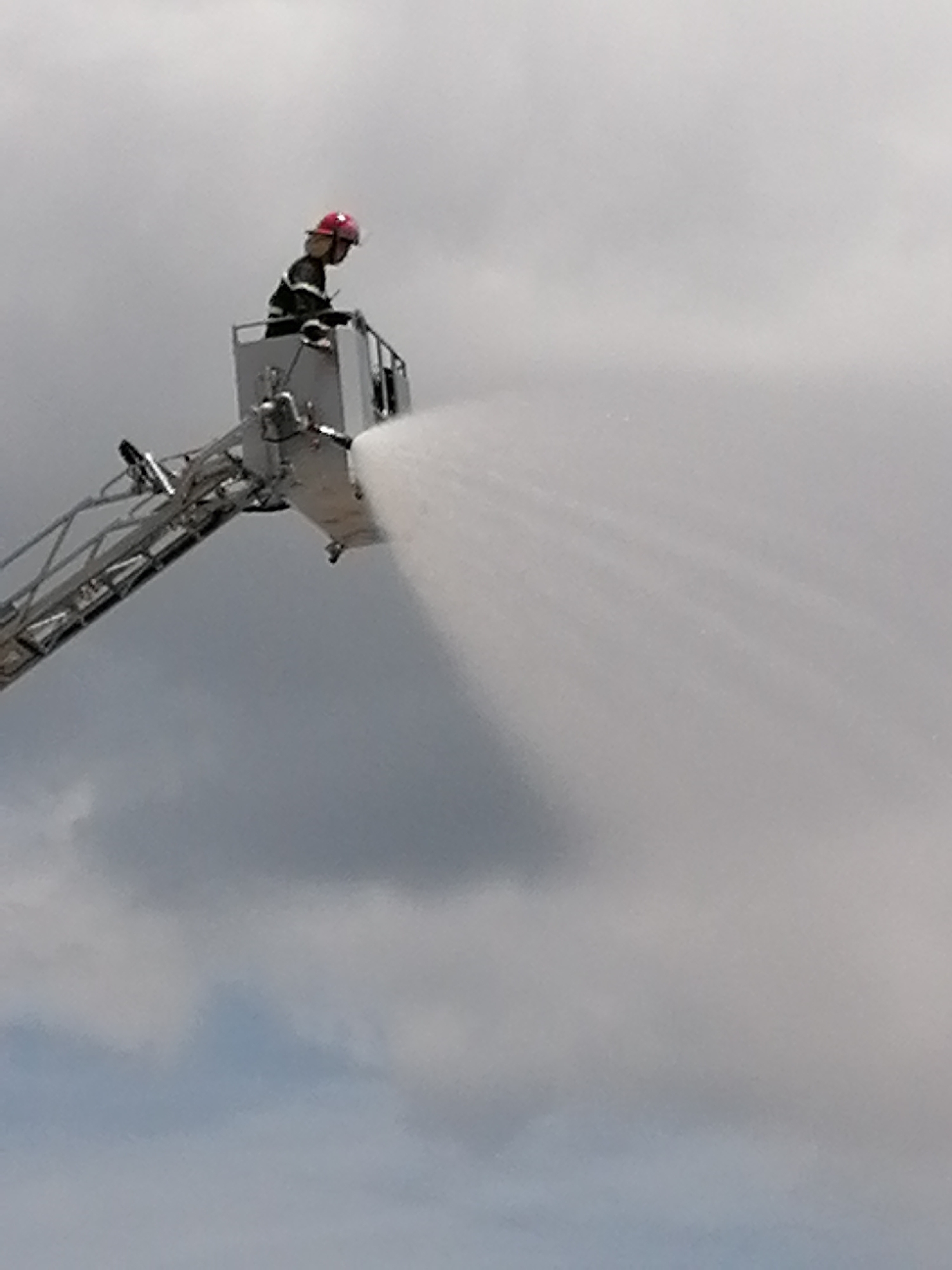 A firefighter, wearing a red helmet and a dark uniform, stands in the basket of an elevated ladder truck, aiming a powerful stream of water. The truck's ladder extends diagonally upward and to the left, with visible metal rungs and support structures. The background consists of a cloudy, overcast sky, with hints of blue peeking through in some areas. There is no identifiable cityscape or specific location visible in this image. The scene captures an action shot of the firefighter operating the water hose.