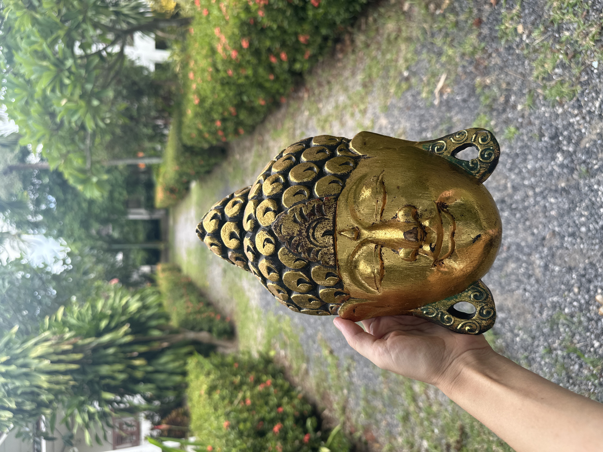 A human hand is holding a golden, intricately carved Buddha mask in the foreground. The mask depicts a serene face with closed eyes and ornate curly hair. The background reveals a garden pathway lined with manicured hedges and red flowers, characteristic of Baan Mae Nam, Thailand. Lush green trees and white buildings are visible further down the path, suggesting a tropical environment. The lighting indicates it is daytime, with soft natural light illuminating the scene.
