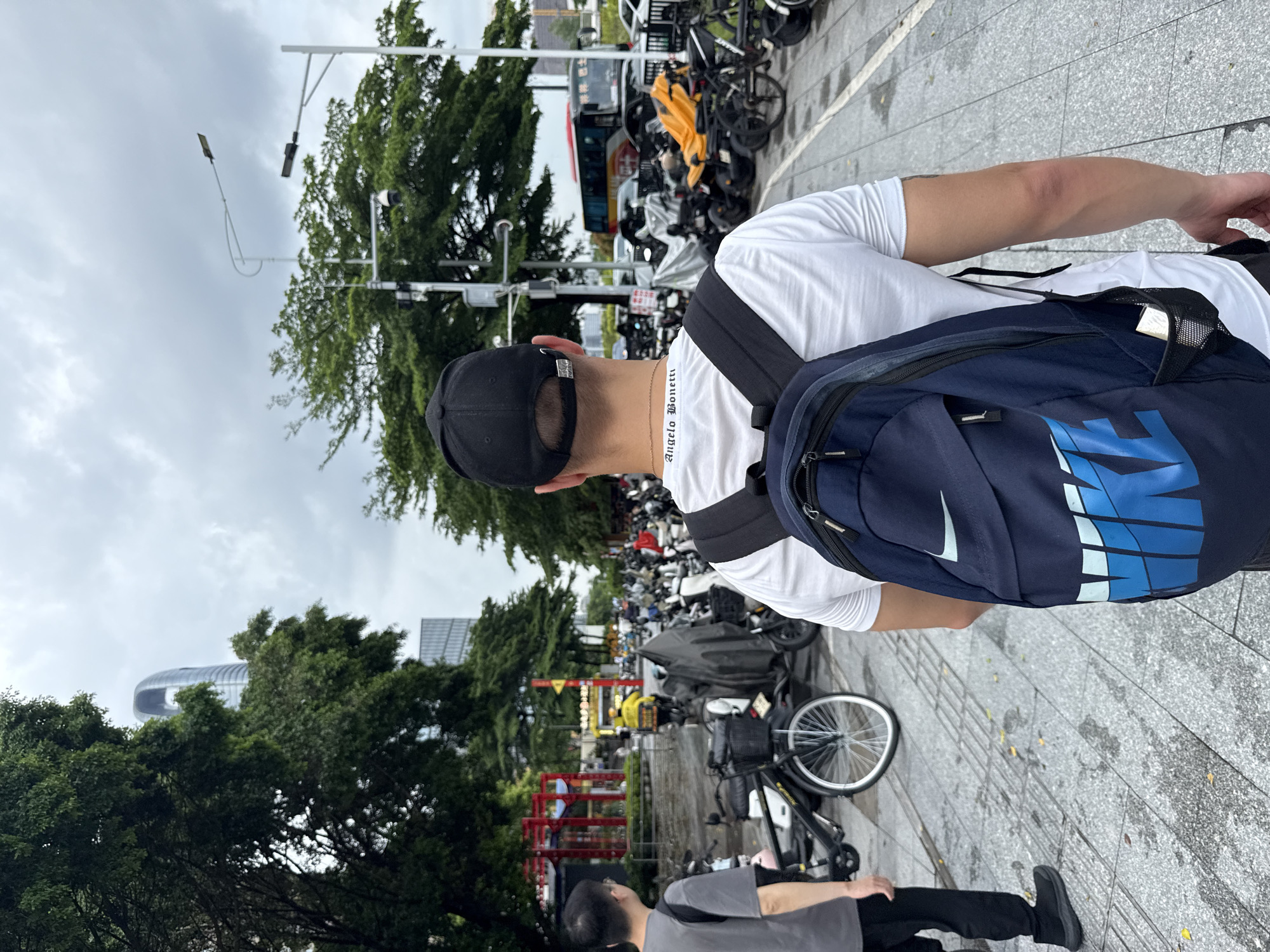 A man wearing a white t-shirt, black baseball cap, and a blue backpack is standing on a wet, paved sidewalk. The backpack has a large blue Nike logo on it. He is looking away from the camera towards a street lined with trees and parked vehicles, including bicycles and scooters. In the background, there is a glimpse of modern architecture and a cloudy sky, suggesting it might be an overcast day. Another person, wearing a grey t-shirt and black pants, is visible further down the sidewalk. The scene appears to be in an urban setting, possibly a public area where people park their vehicles. The text "Angelo Bonetti" is visible on the man's t-shirt. The image is taken in Haizhu District, China.