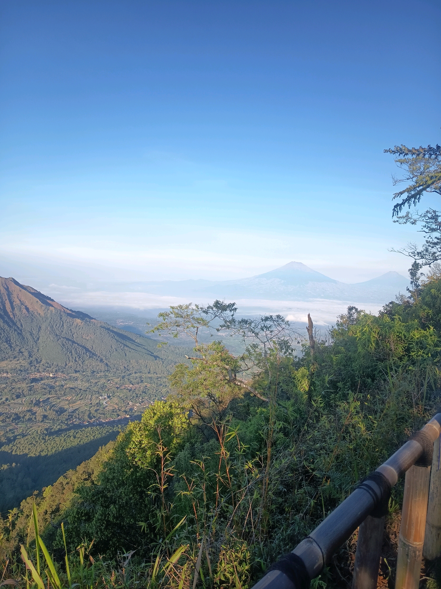 An eye-level, wide shot captures a panoramic view from a mountaintop overlooking a valley filled with mist and distant mountains. The foreground is dominated by lush green foliage and a rustic bamboo railing, suggesting a viewpoint or trail. The landscape stretches out below, with a prominent mountain slope on the left covered in dense trees and a village nestled in the valley floor. In the distance, two conical mountains rise above the mist, one slightly larger and closer than the other. The sky above is a clear, vibrant blue, indicating a bright, sunny day. The scene appears to be located in Keditan, Indonesia, given the mountainous terrain and lush vegetation characteristic of the region.