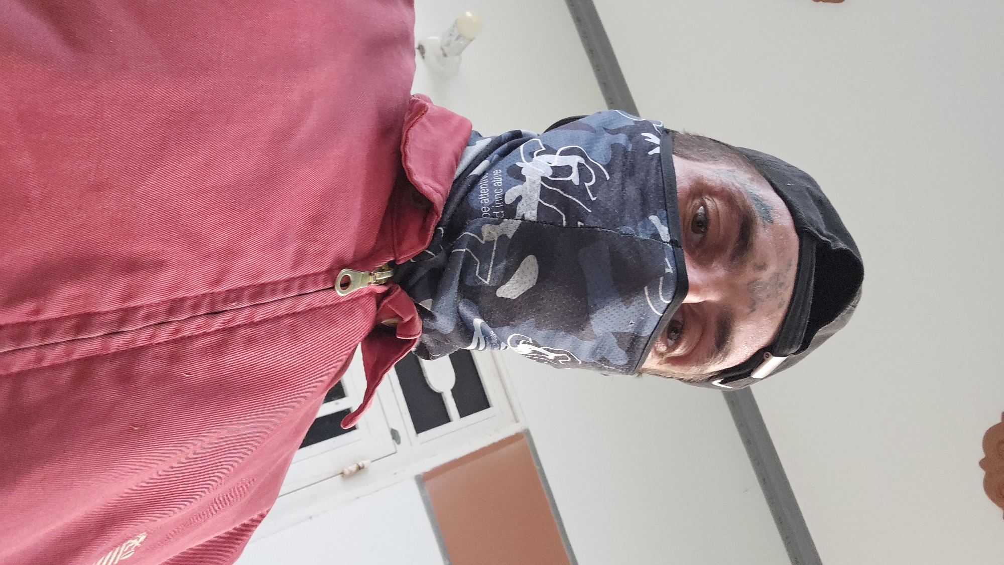 A person wearing a red jacket and a camouflage patterned face covering is seen in a low-angle shot. The person has a black cap, light eyes, and visible tattoos around their eyes. Smudges are visible on their face. A portion of a white window frame and a brown surface are visible in the background. The location is Cam Đức, Vietnam.