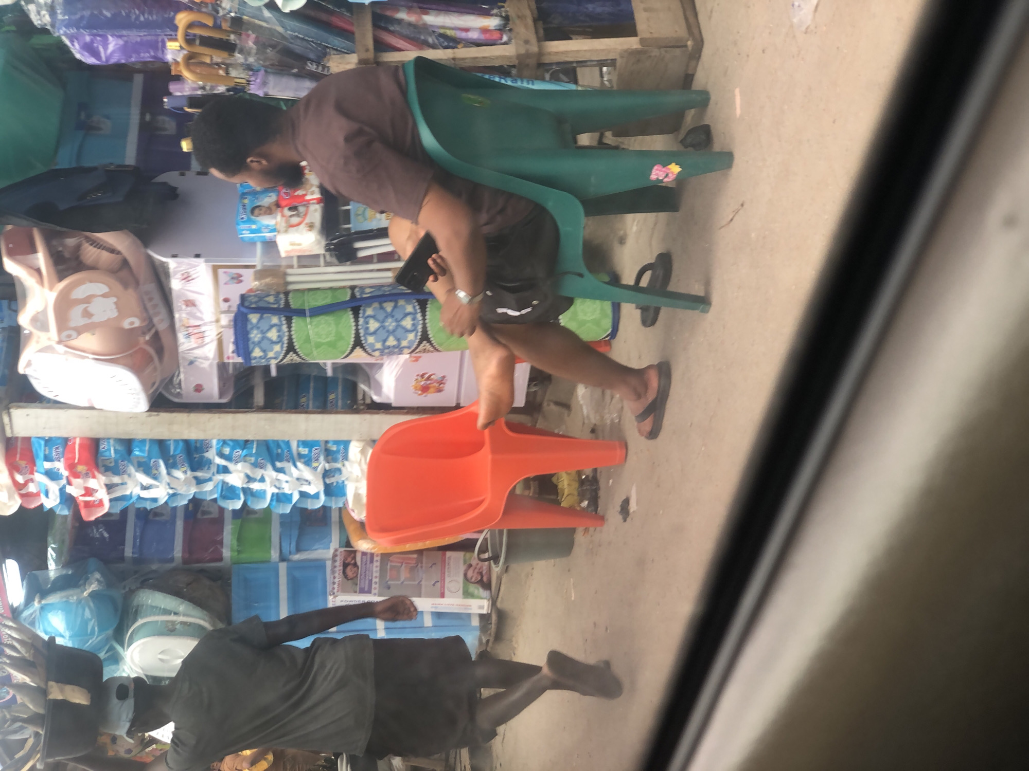 In an outdoor market setting in Lagos, Nigeria, a man walks from left to right, carrying a large black basin filled with fish on his head. He wears a dark grey t-shirt and dark shorts, with dark sandals on his feet. To his right, a second man is seated on a green plastic chair, facing away from the camera and looking down at a black smartphone in his hands. He is wearing a brown short-sleeved shirt and dark shorts, with dark flip-flops on his feet. Next to him, to his left, is an orange plastic chair. The ground is a dirt or sand-like surface. Behind the seated man, there are various market stalls with stacked goods, including plastic containers, umbrellas, and packaged items. The overall scene depicts a typical street market environment.