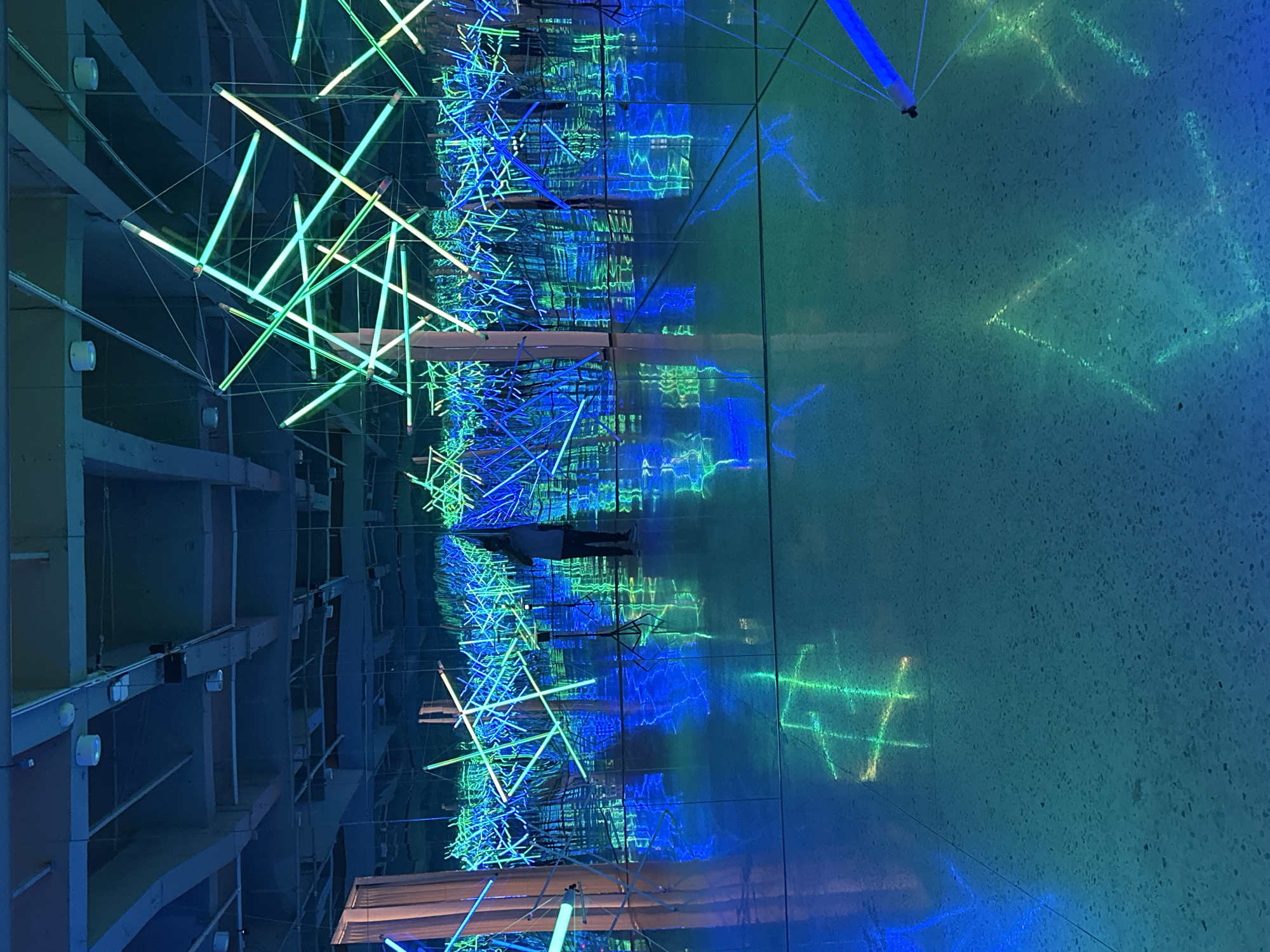 The image presents an abstract, mirrored scene featuring linear lighting fixtures in shades of green and blue, arranged in a seemingly random yet structured pattern. These lights are reflected multiple times, creating an illusion of depth and infinity. A faint silhouette of a person can be discerned amidst the reflected lights, positioned in the center of the perceived corridor. The walls and floor appear reflective, enhancing the effect of an endless tunnel. The color palette is dominated by cool tones, particularly blue and green, creating a futuristic and somewhat ethereal ambiance. The scene seems to be a part of an art installation or architectural feature within a contemporary space. There is no explicit evidence to confirm the image's location in Banan, China.