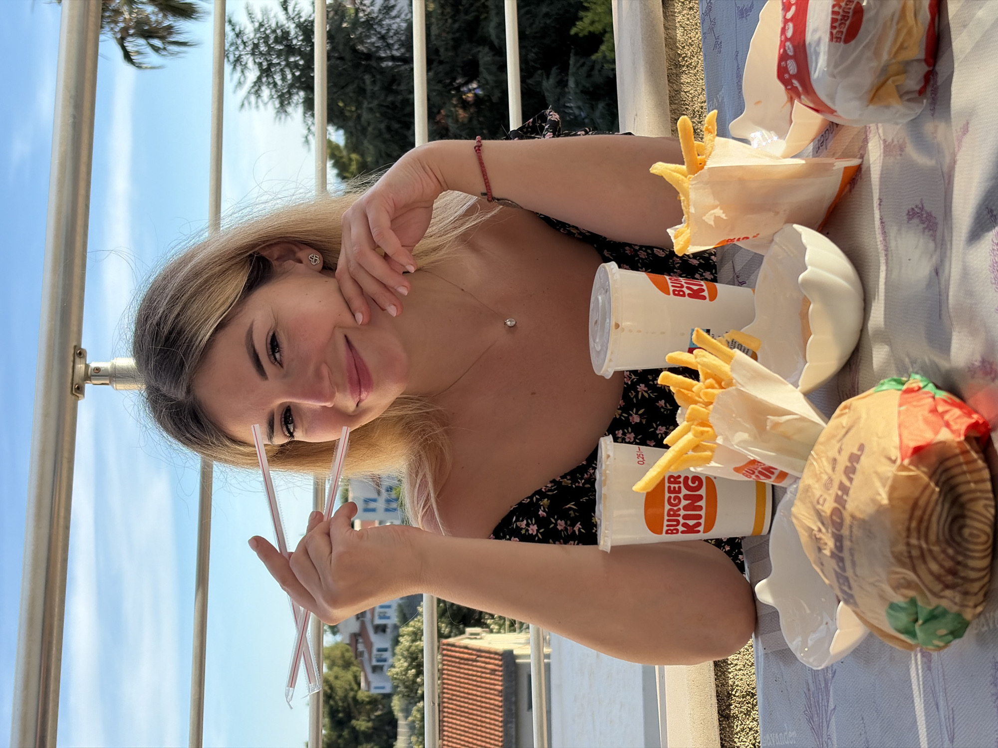 In Budva, Montenegro, a young woman with blonde hair sits at a table outdoors, holding three plastic straws. She is wearing a floral-patterned off-the-shoulder top and a red bracelet. A Burger King meal, including a Whopper burger, french fries in a paper bag, and two drinks, is laid out on the table. The background features a balcony railing, trees, and buildings under a clear blue sky.
