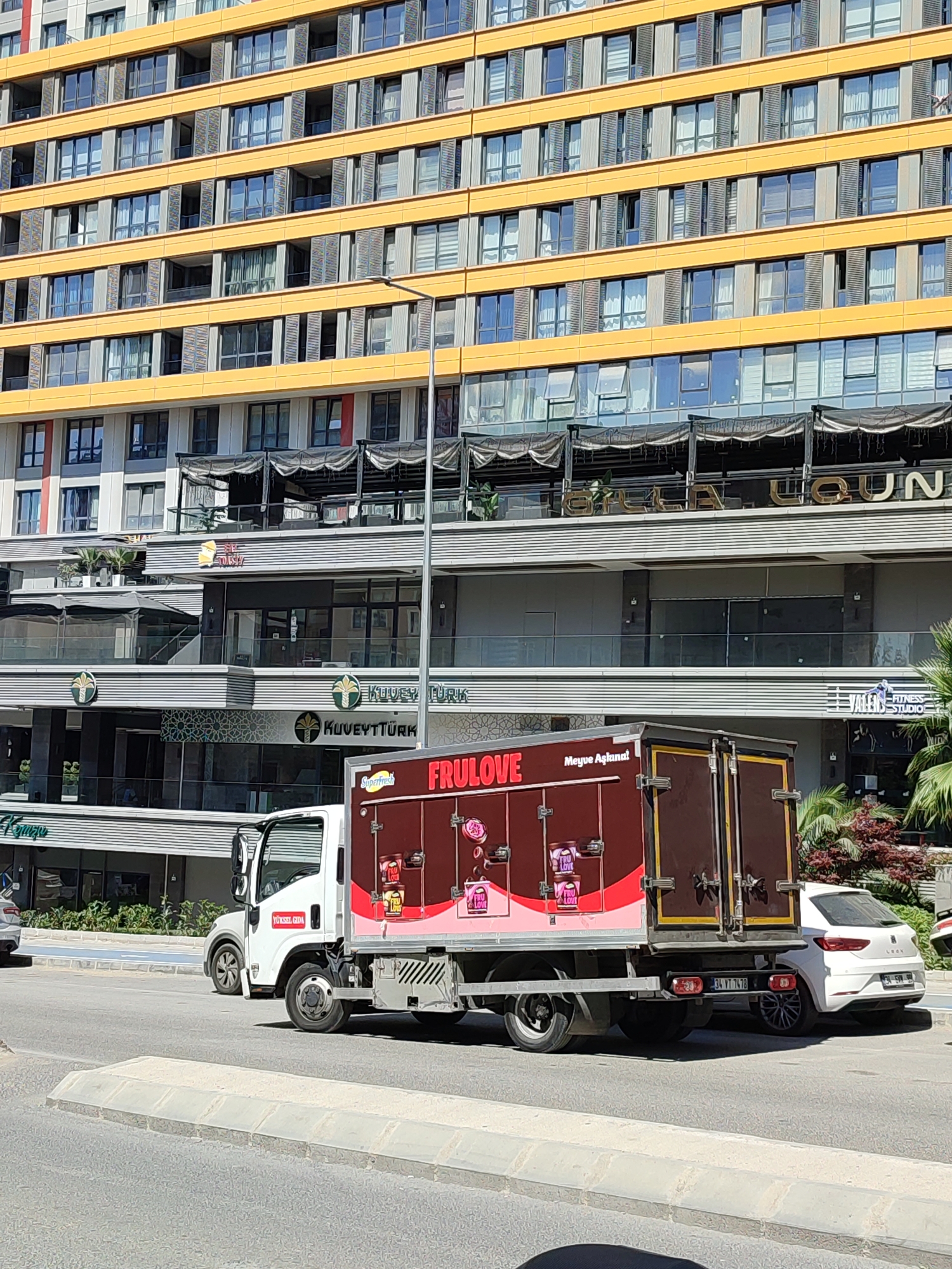 In Kadıköy, Turkey, a white delivery truck with a dark brown cargo box is parked on the right side of a paved road, facing left. The truck has the brand name "FRULOVE" and images of fruit on its cargo box. The truck's cargo box is decorated with images of fruit and the text "FRULOVE" and "Meyve Aşığı". A white car is parked behind the truck. In the background, a modern, multi-story building with yellow and gray accents dominates the scene. The building has numerous windows and balconies, and at street level, there are shops and restaurants with outdoor seating areas. The sky is clear and blue.
