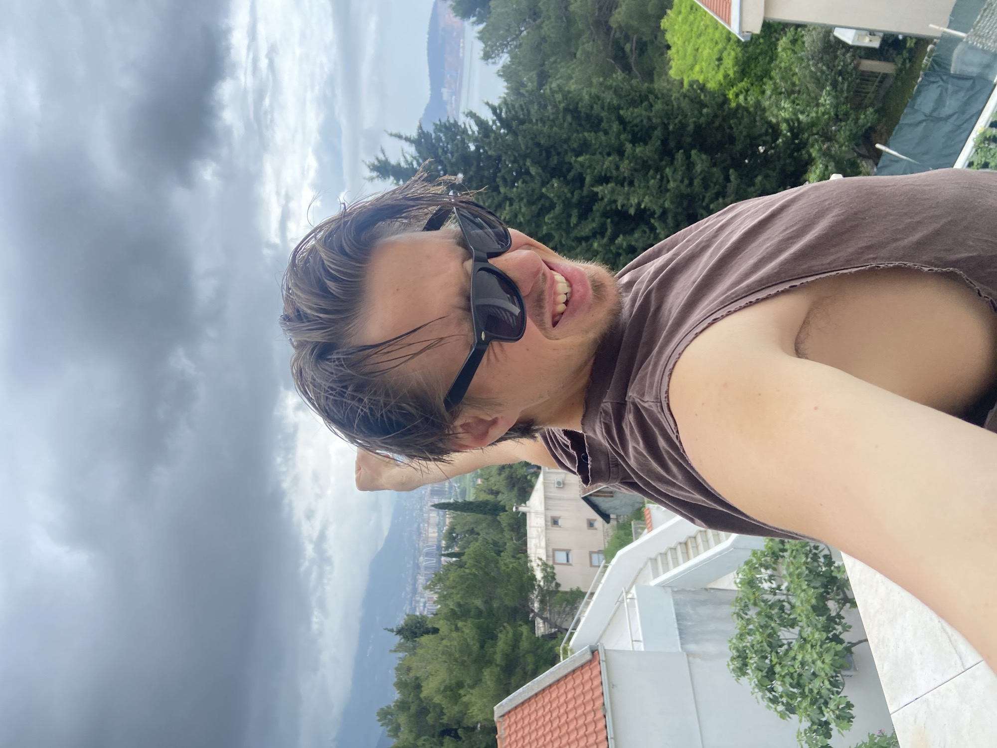 A man with damp, blonde hair and wearing black sunglasses and a brown tank top stands on a balcony and points his right arm towards the horizon. He is smiling widely, showing his teeth. He appears to be facing downwards towards the camera. The background includes a cloudy sky, green trees, a view of the sea, and buildings. The location is likely in the city of Šušanj, Montenegro, as that is the location indicated in the prompt. The balcony features a white railing and red tile roof.