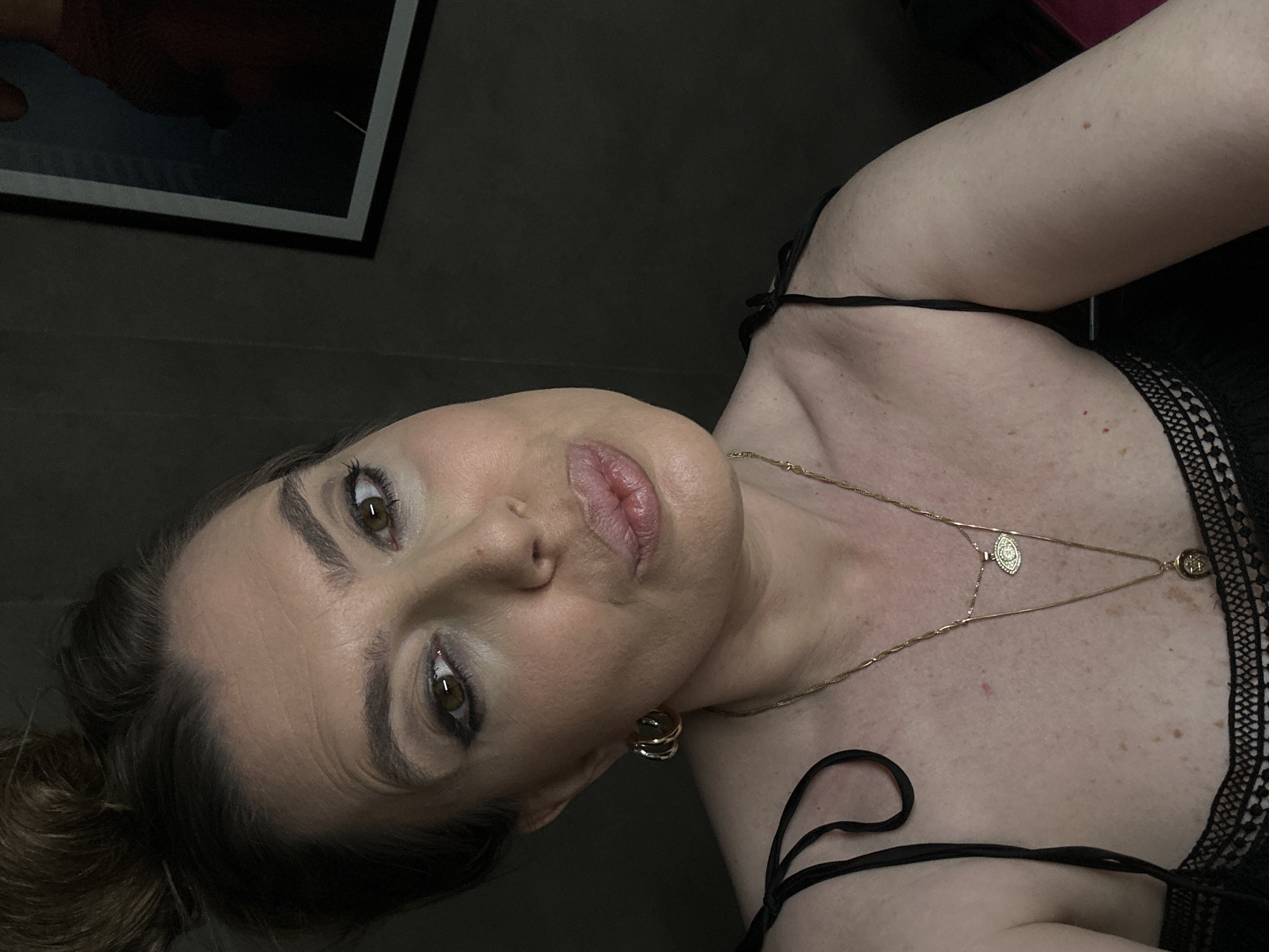 A woman with brown hair and eyes poses indoors, wearing a black lace-trimmed garment and gold jewelry including earrings and a layered necklace. She is positioned against a dark background with a framed artwork partially visible in the upper left corner. The location, Tbilisi, Georgia, is not discernible from the image.