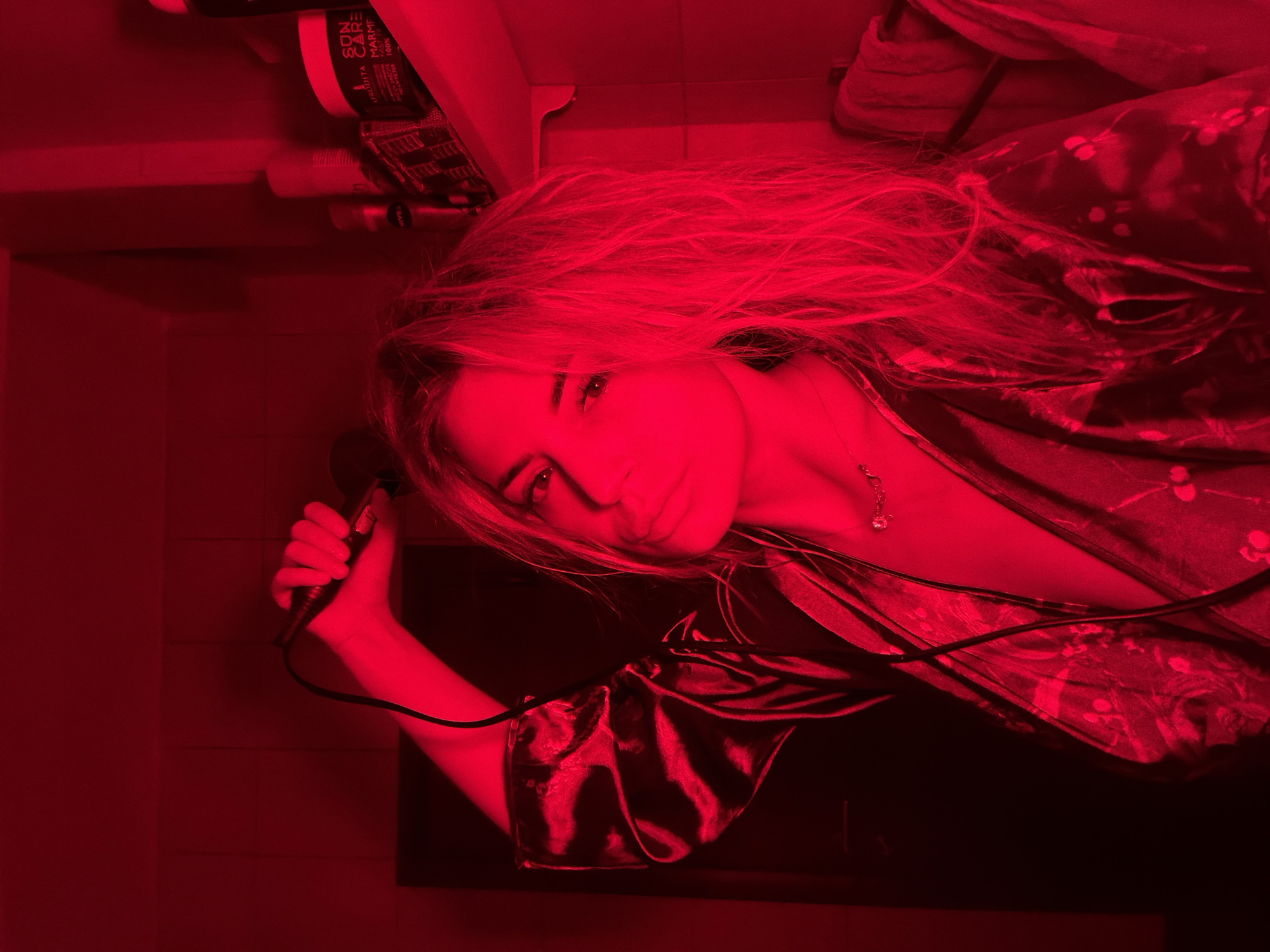 Within a room in Budva, Montenegro, intensely illuminated by a dominant red ambient light, a female subject with long, light-colored hair is depicted. She wears a satin-like, patterned robe with a V-neckline, revealing a delicate necklace. Her right hand is raised above her head, grasping a black, corded hair styling tool, likely a curling iron. She maintains direct eye contact with the viewer, her expression neutral. To her right, a shelf is visible, holding multiple bottles and jars, including one clearly labeled "NIVEA" and another prominent container marked "SUN CARE" and "MARME". A folded white towel or fabric item hangs on the far right. A dark, rectangular panel or cabinet is visible behind her left shoulder. The red lighting casts deep shadows and saturates the entire scene in red tones, obscuring original colors.