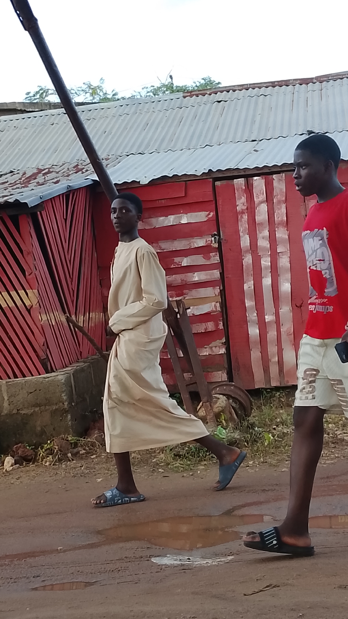 Two young Black males are walking on a dirt road in Adankolo, Nigeria. The male on the left is wearing a long, light-colored robe and dark flip-flops. He is walking away from the camera, looking over his left shoulder. The male on the right is wearing a red t-shirt and white shorts with black stripes. He is walking towards the camera, looking to his left. Behind them is a red wooden building with a corrugated metal roof. There is some vegetation and debris on the ground. The sky is overcast.