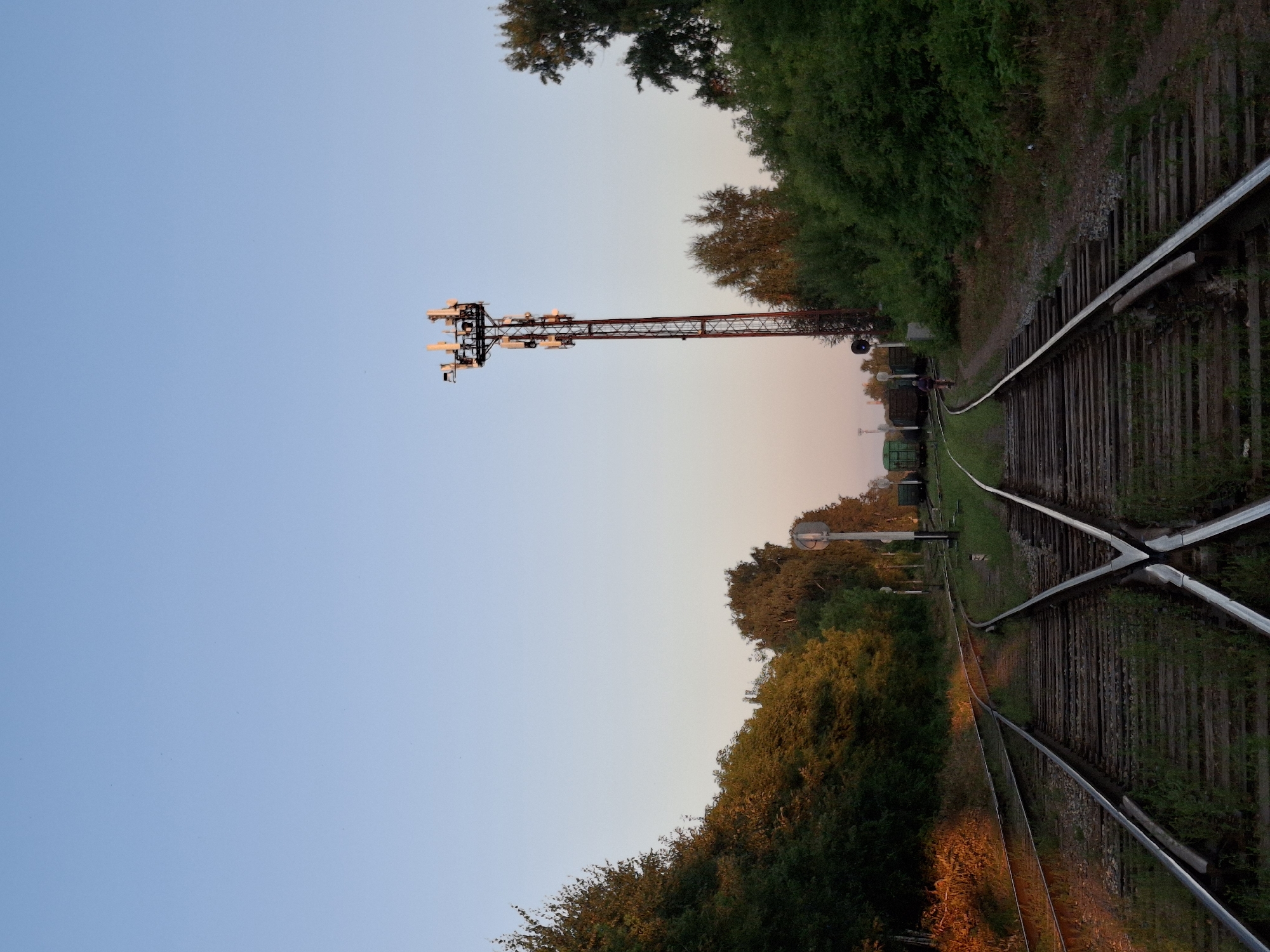 An eye-level, wide-angle shot captures a railway junction with a tall, red metal telecommunications tower to the right, a person on a bicycle riding on the tracks to the right, and green freight cars parked in the distance to the left. The scene is set at dusk with the sky a pale blue and the trees on the left illuminated by the setting sun. The railway tracks are in the foreground, converging and diverging, with grass growing between the wooden sleepers.