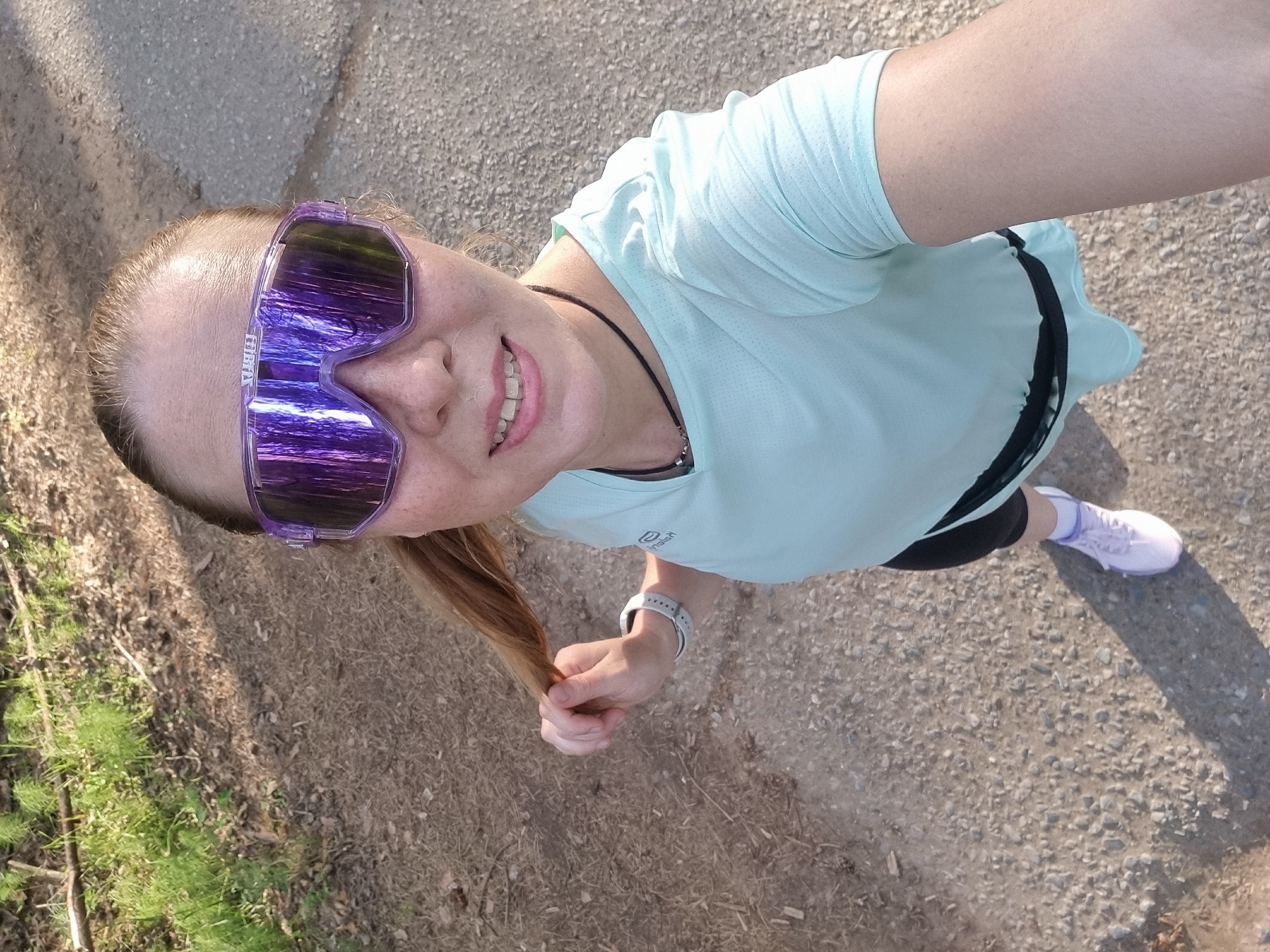 A Caucasian female, appearing to be in her mid-adult years, is depicted in a high-angle outdoor selfie. Her light brown hair is pulled back, and her face shows freckles on the nose and cheeks. She is smiling, revealing her teeth. She wears large, reflective, purple-tinted athletic sunglasses with the brand "100%" visible on the frame bridge. Her clothing includes a light blue short-sleeved athletic t-shirt with a faint logo on the right chest, black athletic shorts worn beneath, and light purple athletic shoes with white laces and socks. A black cord necklace with a metallic bead is visible around her neck. On her left wrist, she wears a white sports watch. Her left hand is actively holding a section of her ponytail. The ground beneath her is an unpaved path composed of light-colored gravel and dirt, with areas of green grass, dried leaves, and small branches visible along the left side and in the background. A distinct shadow is cast on the ground to her right, indicating bright overhead sunlight. The location is in the city of Chaikovsky, Russia.