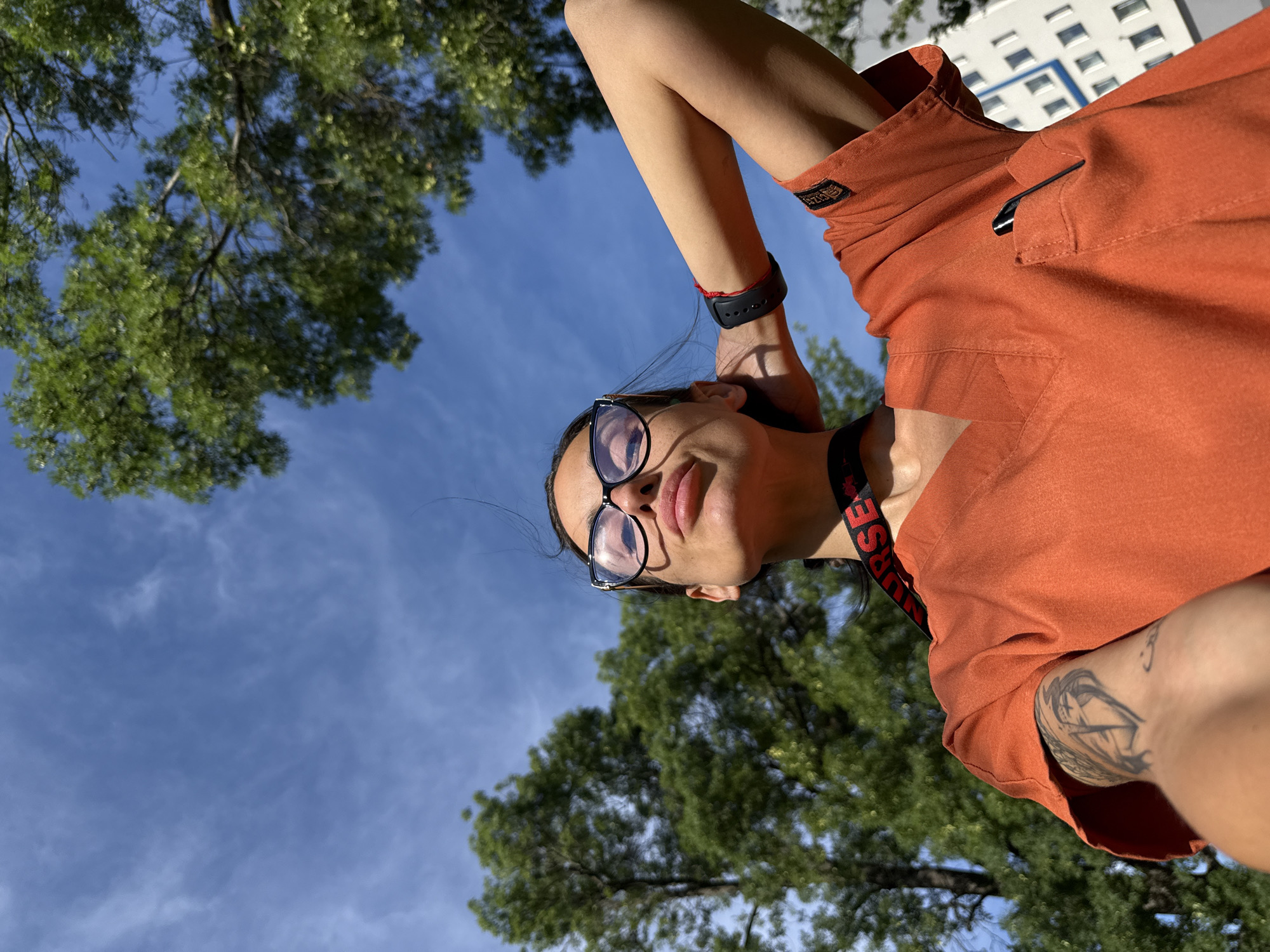 A young woman wearing an orange scrub top and black-rimmed glasses poses for a selfie outdoors. She is looking up at the sky with her eyes closed and her lips pursed. Her right arm is raised behind her head, and she is wearing a black fitness tracker on her left wrist and a red string bracelet on her right wrist. A black lanyard with the word "NURSE" in red lettering is around her neck. A tattoo is visible on her left arm. The background shows a bright blue sky with scattered clouds, green trees, and a white building. The setting is likely a public area, possibly near a hospital or medical facility, given the woman's attire. Without additional context, the specific location within the city of Pleven, Bulgaria, cannot be determined.