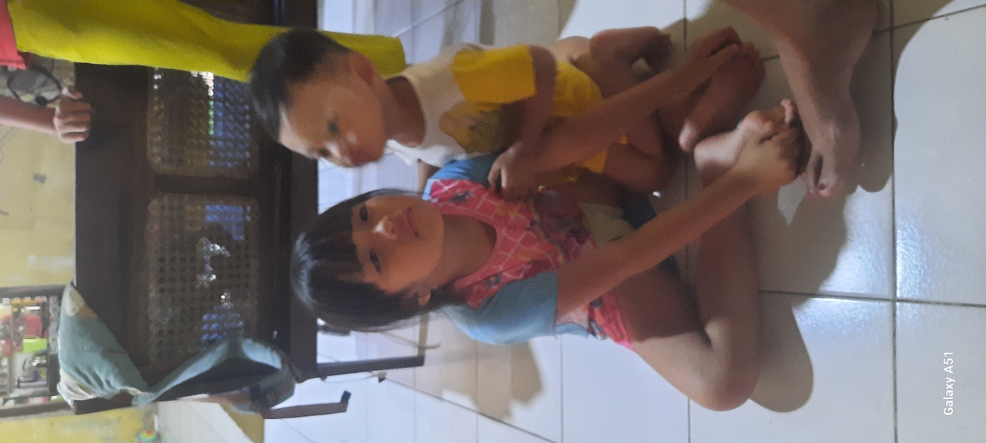 Two young children are sitting on a tiled floor. The child on the left, a girl, has dark hair and is wearing a blue and pink patterned t-shirt and red shorts. She is looking forward with a slightly bored expression. The child on the right, a boy, is facing away from the camera and appears to be leaning towards the girl. He is wearing a yellow and white shirt. Behind the children is a dark-colored piece of furniture with a woven or perforated surface, possibly a cabinet or bench. A light-colored blanket or cloth is draped over the furniture. The background is blurred but suggests an indoor setting with hints of kitchen elements. The lighting appears to be artificial. The lower portion of the image shows the legs and feet of the children, with shadows cast on the tiled floor. The text "Galaxy A51" is visible in the bottom left corner. The scene is located in Ploso, Indonesia.