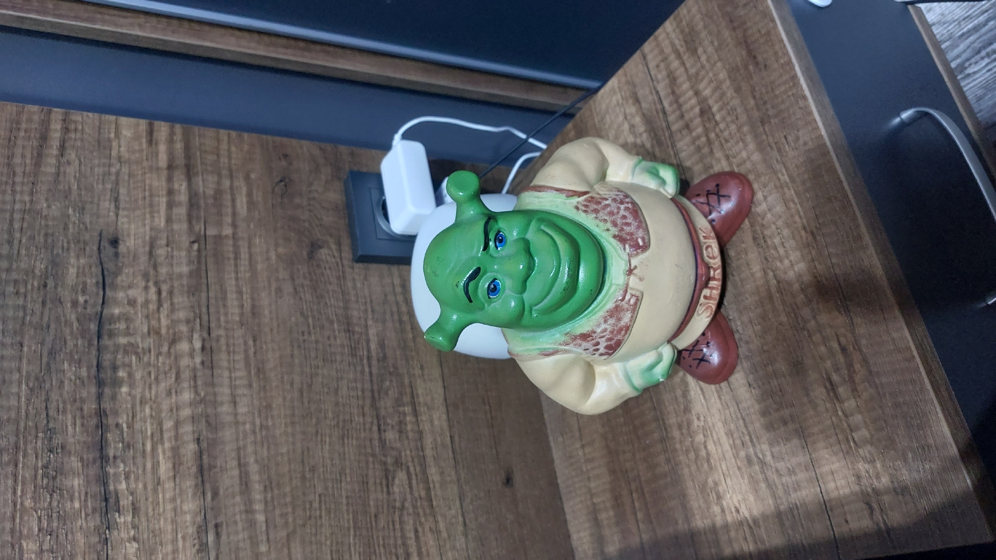 A Shrek figurine rests on a wooden surface, appearing to be a table or shelf. The figurine depicts Shrek's head and upper body, rendered in vibrant green for his skin and a lighter, beige color for his tunic, which has reddish-brown detailing. His blue eyes are open, and he sports a wide, friendly smile. The figurine is oriented sideways in the image, suggesting it might be lying down or the photo was taken at an angle.
Behind the Shrek figurine, a white power adapter is plugged into a black wall socket or power strip, with white cables extending from it. The wooden surface beneath the figurine features a visible wood grain pattern with varying shades of brown. In the background, a dark blue or black surface, possibly another piece of furniture or a wall panel, is visible. On the figurine's right foot, the word "SHREK" is clearly visible in raised letters. The scene is indoors, likely under artificial lighting, and there is no activity taking place beyond the still arrangement of objects. The location context for this scene is Kostandovo, Bulgaria.