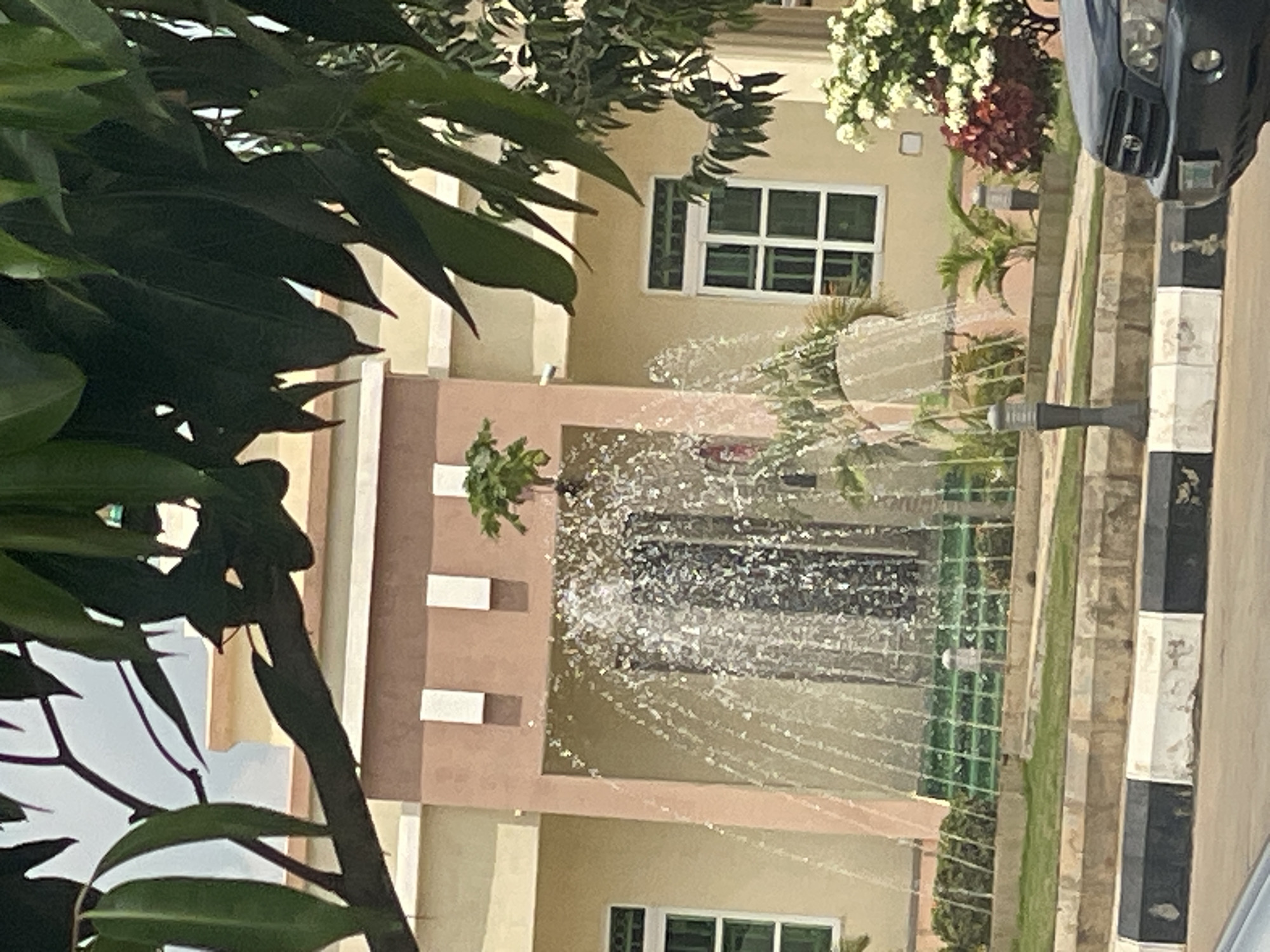 The image depicts a fountain in front of a cream-colored building with pink and brown accents. The building features several windows and is partially obscured by the branches and leaves of a large tree in the foreground. Water from the fountain cascades down, creating a shimmering effect. In front of the fountain is a tiered garden area with green grass and some shrubs. A black car is visible on the right side of the image, parked near a black and white striped barrier. The overall scene appears to be an outdoor residential or commercial area.