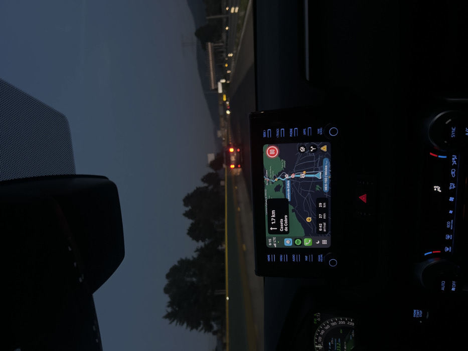 Inside a vehicle, likely at dusk, a navigation screen displays a route with a distance of 1.7 km to "Caseta de Cobro." The estimated arrival time is 6:42, with 27 minutes remaining and 29 km to travel. The speedometer is visible, and dashboard controls for climate and audio are present. The driver is on a highway with other vehicles, as seen through the windshield. The dashboard also features buttons for track, phone, and apps.