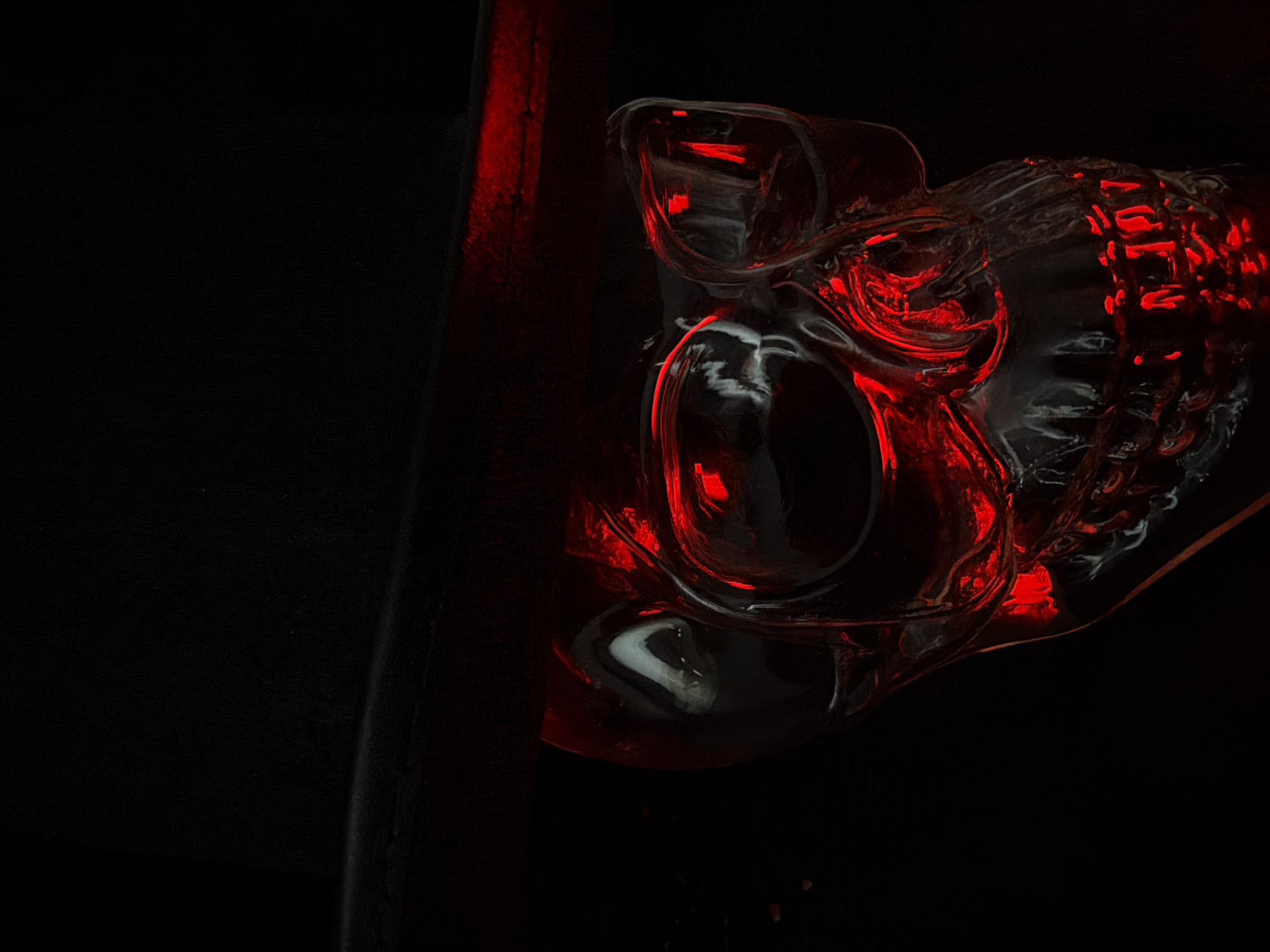 A close-up, low-angle shot shows the top portion of a clear glass skull illuminated from within by a bright red light, casting a sinister glow. The skull is positioned beneath a black top hat, with the brim of the hat casting a shadow over the upper part of the skull. The background is entirely black, creating a stark contrast with the illuminated skull and hat. The glass of the skull is highly reflective, with highlights and shadows playing across its surface, giving it a wet or eerie appearance. The red light appears to emanate from within the eye sockets and the mouth of the skull. The texture of the hat appears to be matte and slightly worn. The overall impression is one of Halloween or gothic macabre. The location is unknown.