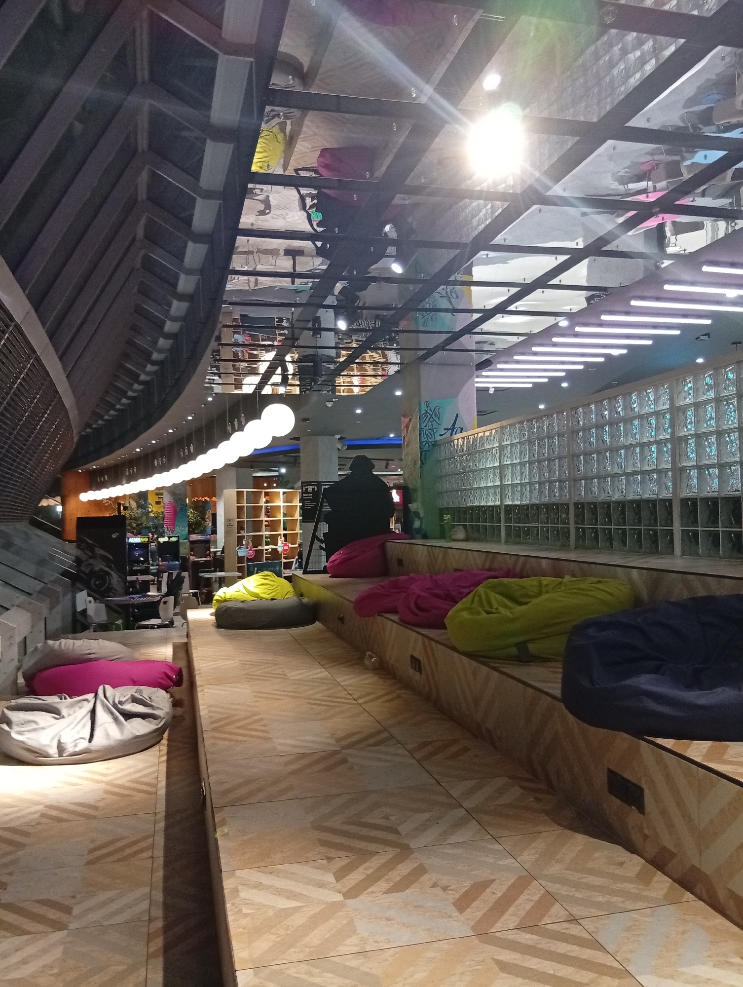 The image depicts an interior scene with a contemporary design, likely a public space or a lounge area. The foreground is dominated by a sloped wooden floor with a geometric pattern, interspersed with several large beanbag chairs in various colors: grey, pink, yellow, and navy blue. These chairs are arranged on stepped wooden platforms that rise towards the background.
In the mid-ground, to the left, is an area with what appears to be arcade game machines, suggesting a recreational or entertainment venue. A person, silhouetted and wearing dark clothing, is visible standing in the mid-ground, facing away from the camera, possibly observing something in the distance or interacting with the games.
The background features a wall constructed of glass blocks, with a segmented wooden paneling below. Above, the ceiling is a complex structure with exposed metal beams and a reflective surface, possibly mirrored panels or polished metal, which reflects the overhead lighting. A long string of large, spherical white lights hangs from the ceiling in the mid-left portion of the image. Linear fluorescent lights are also visible along the ceiling in the upper right. The overall lighting is bright, with a strong glare from an overhead light source.
The scene is clean and orderly, with no visible signs of disarray or immediate activity beyond the presence of the beanbags and the single standing figure. The environment suggests a modern and comfortable space designed for relaxation or entertainment. The presence of arcade games and the general ambiance might indicate a space within a mall, entertainment complex, or a trendy cafe.