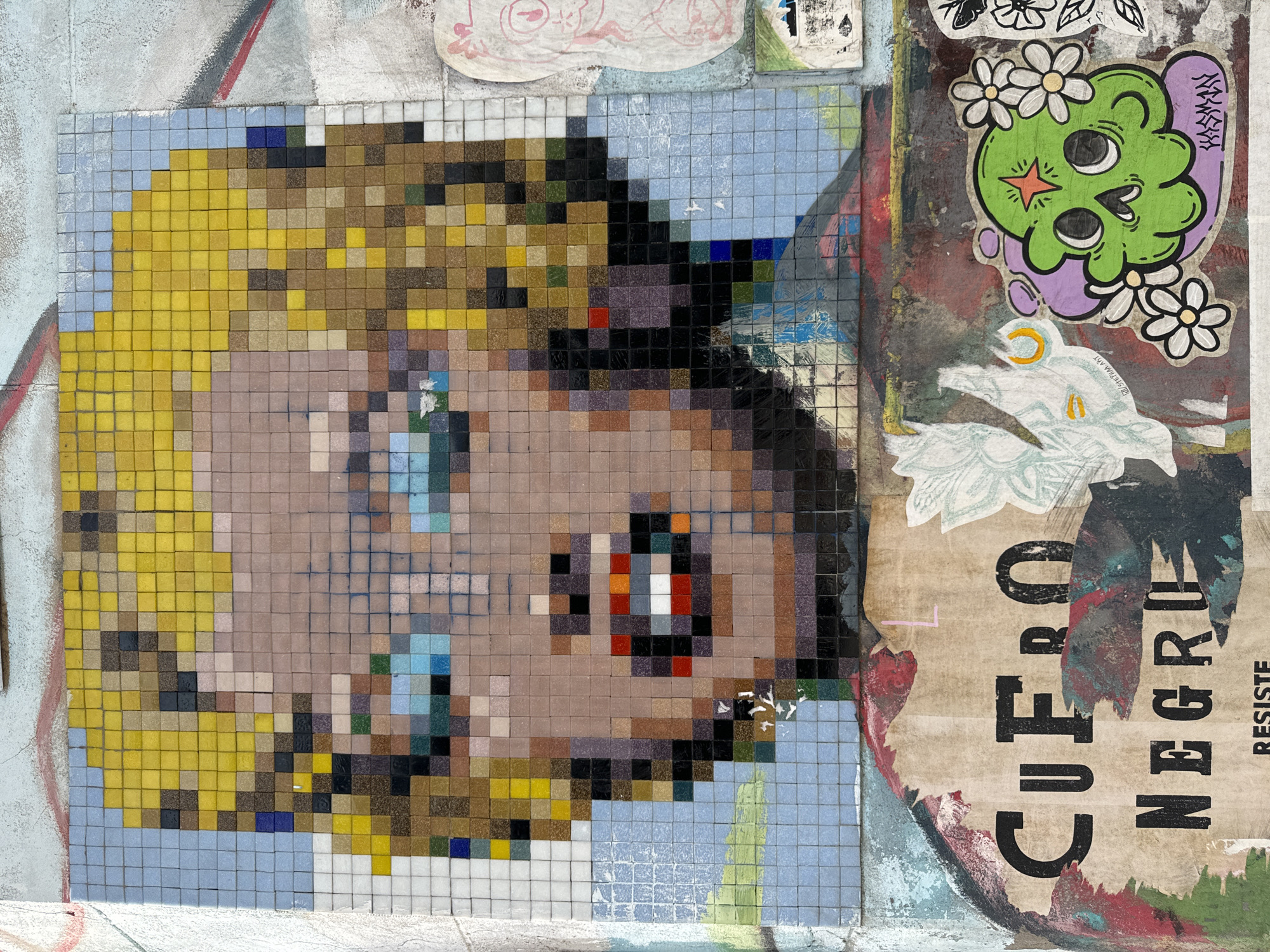 The image captures a section of urban street art in Buenos Aires, Argentina. A mosaic depicting a pixelated, blond-haired person's face dominates the left side, constructed from small square tiles in varying shades of yellow, brown, beige, blue, and black. The person's face appears to be looking to the right.
To the right of the mosaic are several layers of peeling posters and stickers. One sticker features a green cartoonish figure with a purple outline and white flower accents. Another poster is partially ripped, revealing the words "CUERPO NEGRO" in large black letters, with the word "RESISTE" visible further down.
The surrounding wall is painted with various colors and textures, adding to the layered and textured nature of the street art composition.