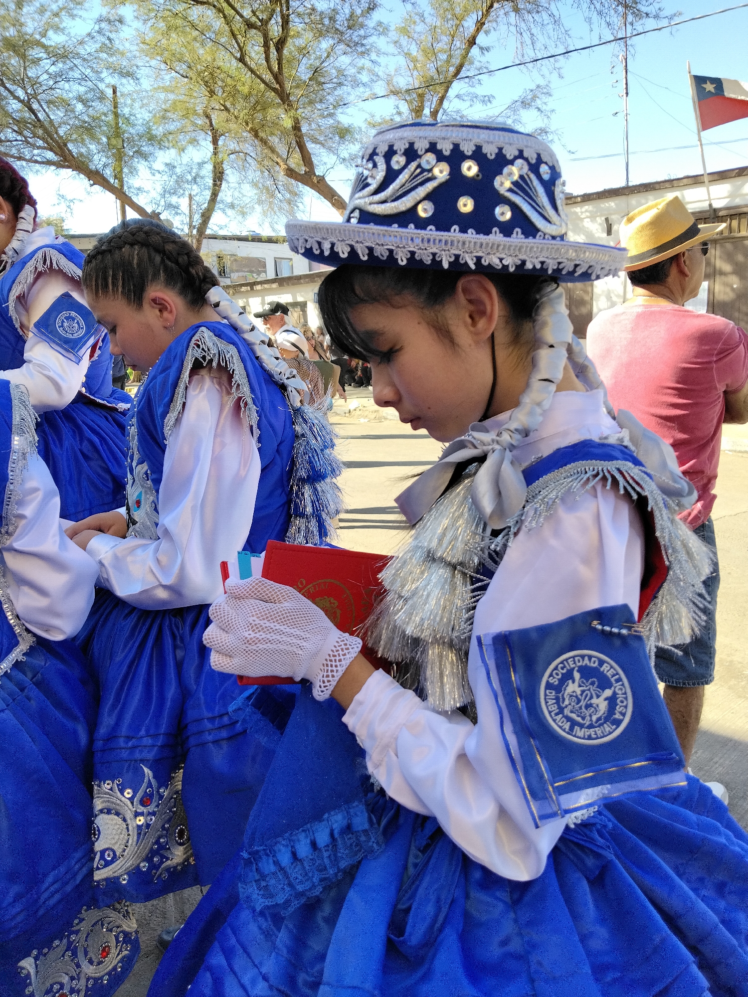 In Pozo Almonte, Chile, under a clear sky, two young individuals are dressed in traditional blue and white ceremonial attire. The focus is on the rightmost individual, whose face is partially obscured as they look down, holding a red book and a white lacy glove in their gloved hand. Their elaborate blue headdress is adorned with white beadwork and decorative silver accents. The costume features a white blouse with puffed sleeves and a blue, intricately embroidered skirt. The individual on the left, also in similar attire, is partially visible, their braided hair adorned with white ribbons. In the background, a person wearing a yellow hat and a pink shirt is visible, as is the Chilean flag flying in the distance. The setting appears to be an outdoor event or procession.