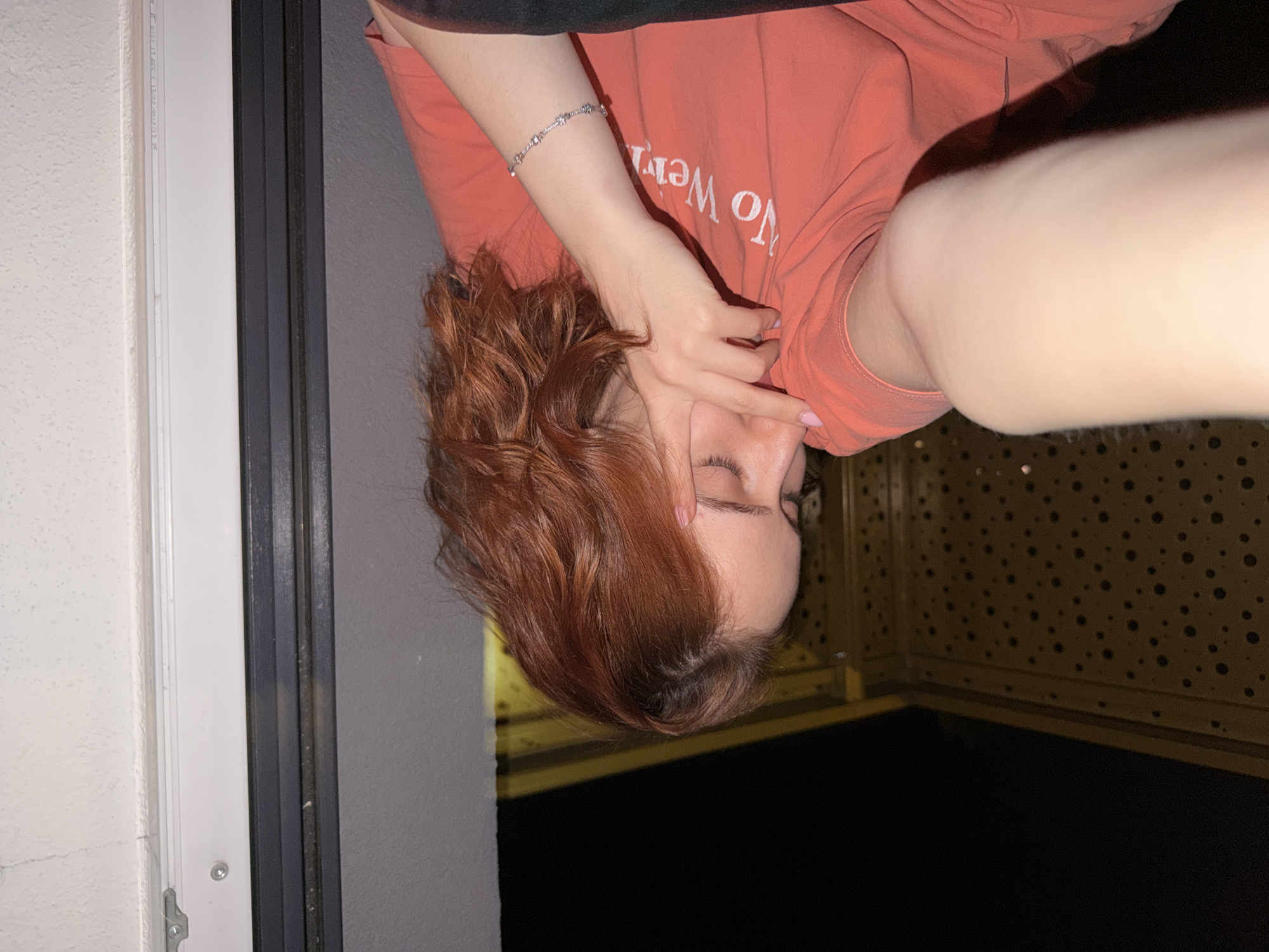 The image shows a young woman with reddish-brown hair, wearing an orange t-shirt with white lettering that reads "No Worries", and a silver bracelet on her left wrist. She is holding her right hand up to her face, covering her eyes and forehead. The background appears to be a balcony or outdoor space at night, with a dark sky, a yellow railing with a pattern of holes, and a white wall. The lighting suggests an indoor or semi-enclosed area with artificial light. There is no discernible evidence or context to suggest the location is in Strasbourg, France. The scene depicts a private moment, possibly a selfie, with the subject looking down and away from the camera.