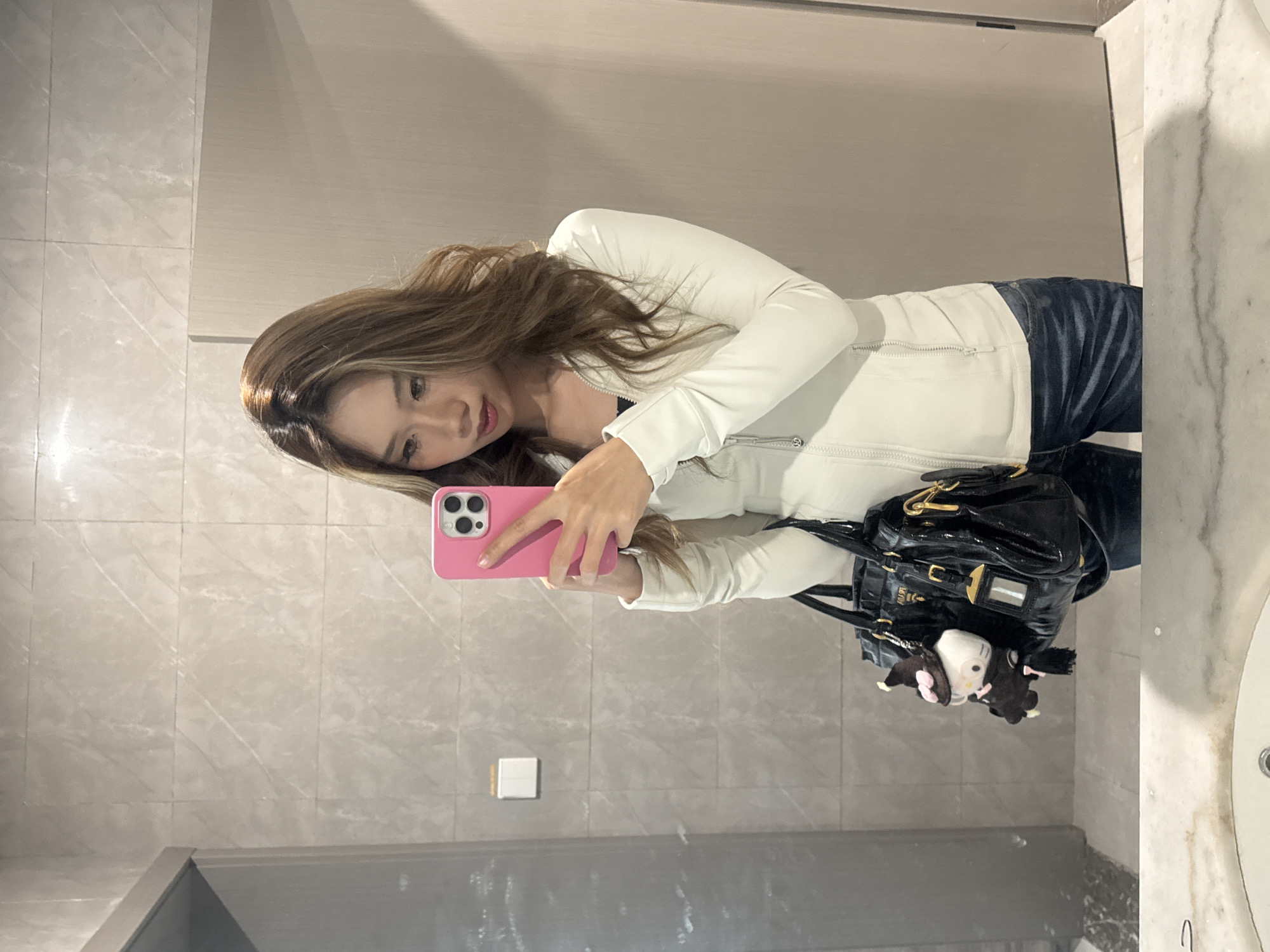 A young woman is taking a selfie in a bathroom. She is holding a pink smartphone and looking at the camera. She is wearing a white jacket and blue jeans. A black purse hangs from her shoulder. The bathroom has a marble countertop, tiled walls, and a sink. It is possible the location is in Nan'an District, China.