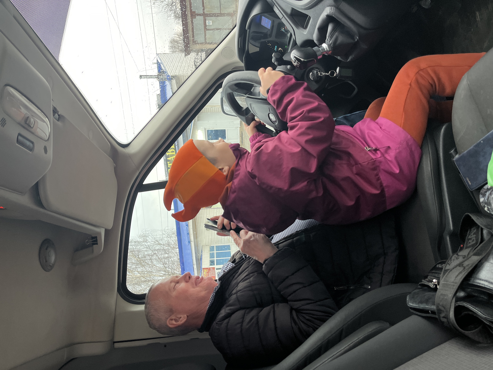 A fair-skinned adult male sits in the passenger seat of a vehicle, appearing to look at and/or interact with a mobile phone. The man is middle-aged with short, gray hair and is wearing a black, puffy jacket. He appears to be smiling.
A child, wearing an orange and yellow hat and a magenta jacket, is positioned across the adult male's lap and is reaching for the steering wheel. The child is wearing orange leggings. The background suggests they are in a parking lot, near a building.
The inside of the vehicle is visible, including the dashboard and other internal features. Based on the exterior signage visible through the windshield, the location of this scene is Noviy, Russia.