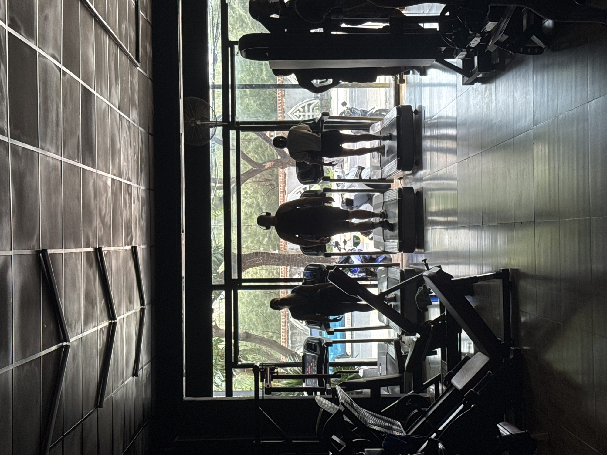 The image shows a gym in Đà Nẵng, Vietnam. Three people are using treadmills near a large window overlooking a street with trees and buildings. Other gym equipment is visible, including weight machines. The room is dimly lit, with a grid-patterned wall to the left. The people are silhouetted against the bright light from the window. A fan is mounted on the wall near the window.