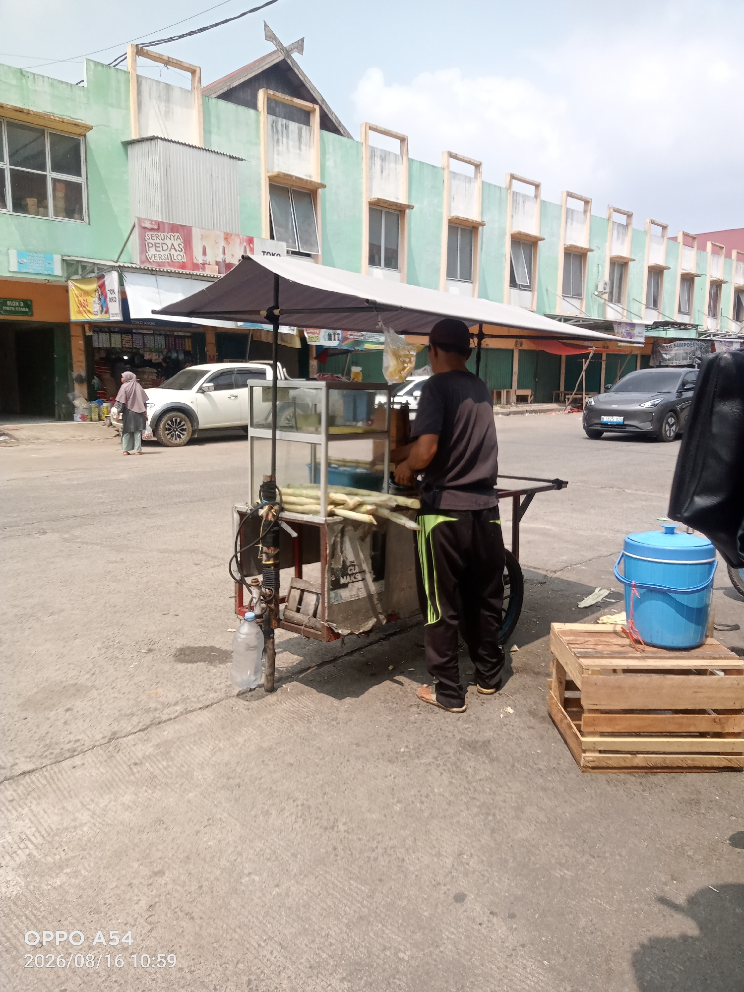 The image depicts a street scene in Jambi, Indonesia, likely in a market or commercial area. In the foreground, a man is operating a mobile sugarcane juice stand. He is seen from the back, wearing a dark t-shirt and black pants with neon green stripes. He is standing next to a cart with a glass display case, where sugarcane stalks are visible. The cart has a black umbrella providing shade. To the right of the cart, a blue bucket sits on a wooden pallet. In the background, a white pickup truck is parked, and a woman in a headscarf is walking away from it. Beyond the street, there is a row of multi-story buildings painted in light green and cream colors, with various shops and awnings visible. The sky is bright and appears to be a clear day. The overall environment suggests a bustling commercial street in Jambi.