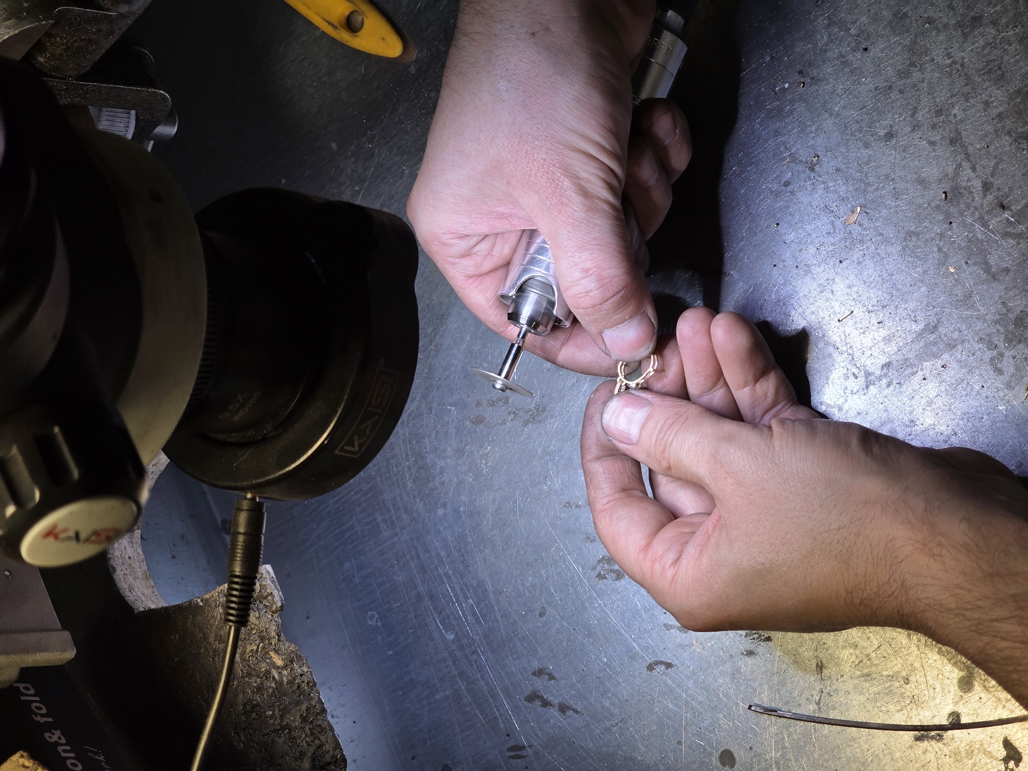 A close-up, overhead shot shows a person's hands working on a piece of jewelry under a microscope. The workbench is a metallic surface with scratches and a few small debris pieces. A rotary tool with a small cutting disc is held in the right hand, and it is actively being used on a gold-colored jewelry piece being held by the left thumb and index finger. The jewelry piece appears to be an ornate setting with prongs, possibly for a stone. To the left, a thin wire or rod is visible on the workbench. The background shows part of a microscope with a dark, cylindrical base and a silver-toned arm. A yellow tool is also partially visible on the right side of the image. The scene is likely indoors, in a workshop environment, possibly related to jewelry repair or creation. The location in Kazan, Russia, cannot be determined from the visual information in the image.